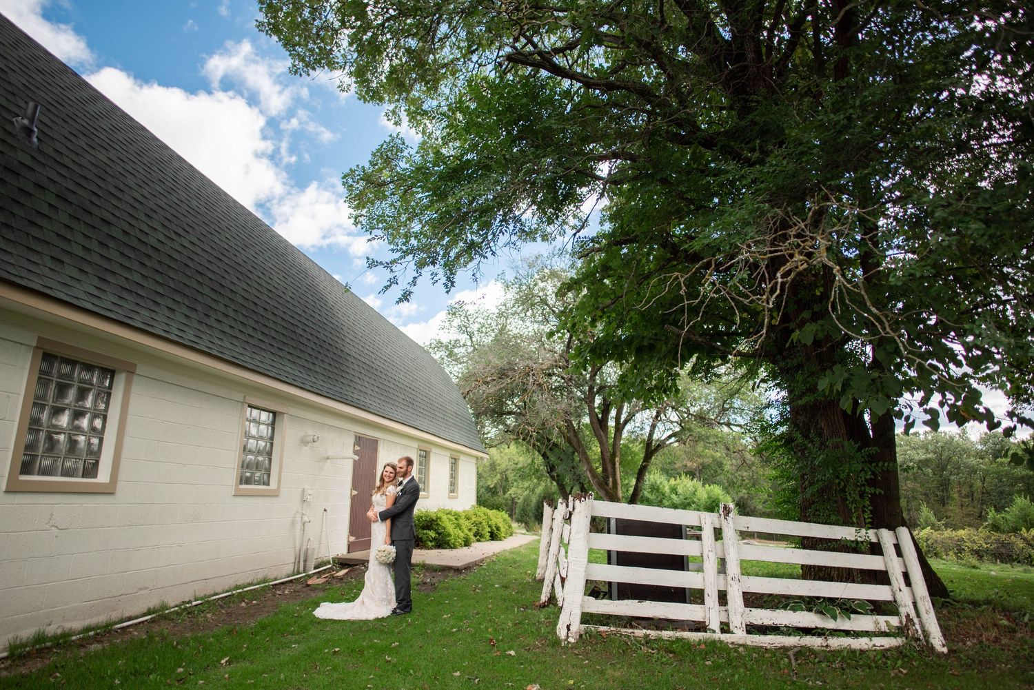 A couple celebrating their wedding outside a charming white country house with white picket fence and lush green trees.