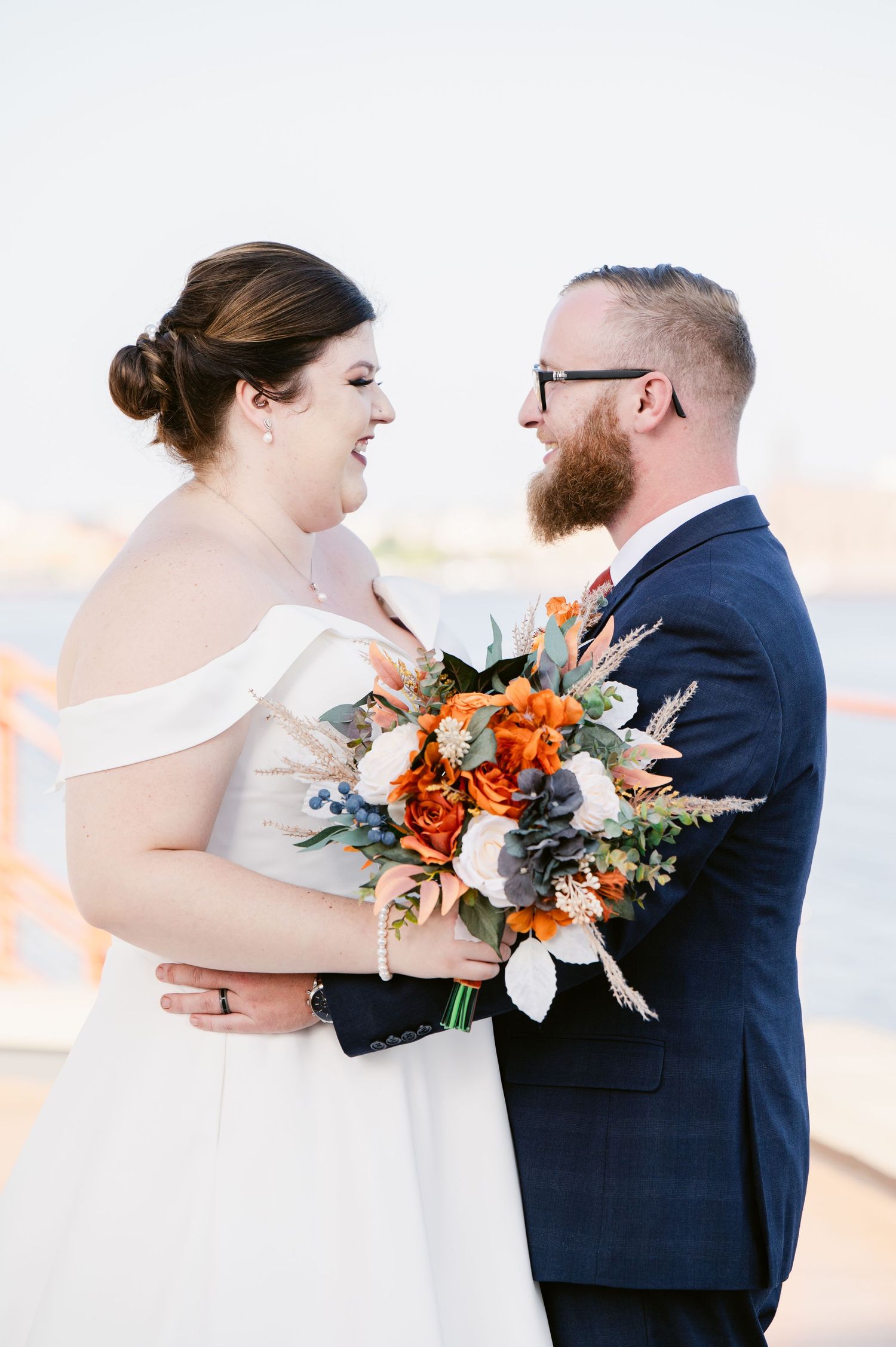 Pier B Duluth Wedding Wedding couple shares romantic moments outdoors with orange floral bouquet in white dress and navy suit.
