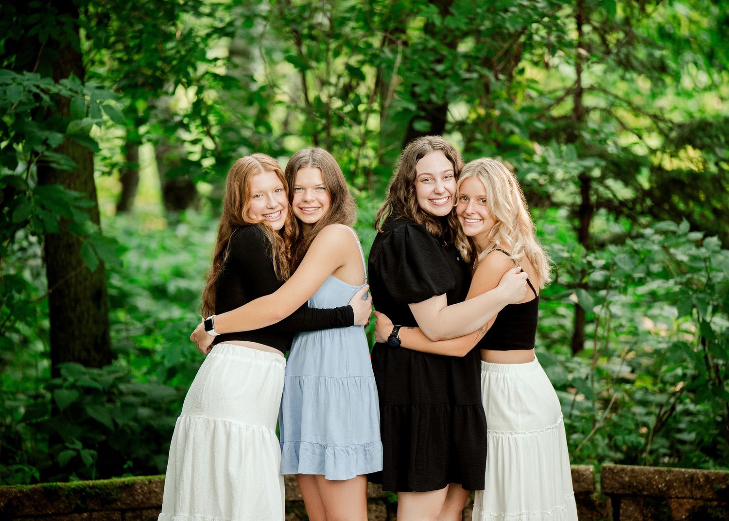 A group of friends in white and black dresses pose together in a lush green forest setting.
