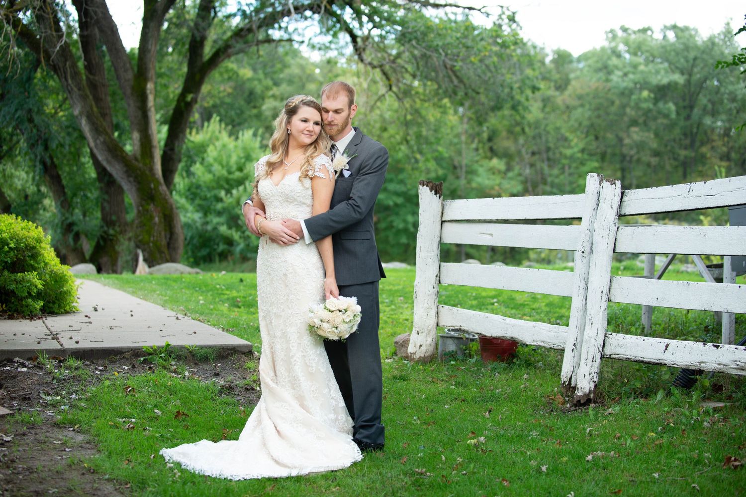 A couple in wedding attire embrace near a white fence on a lush green garden path. Rum River Barn, Hillman MN.