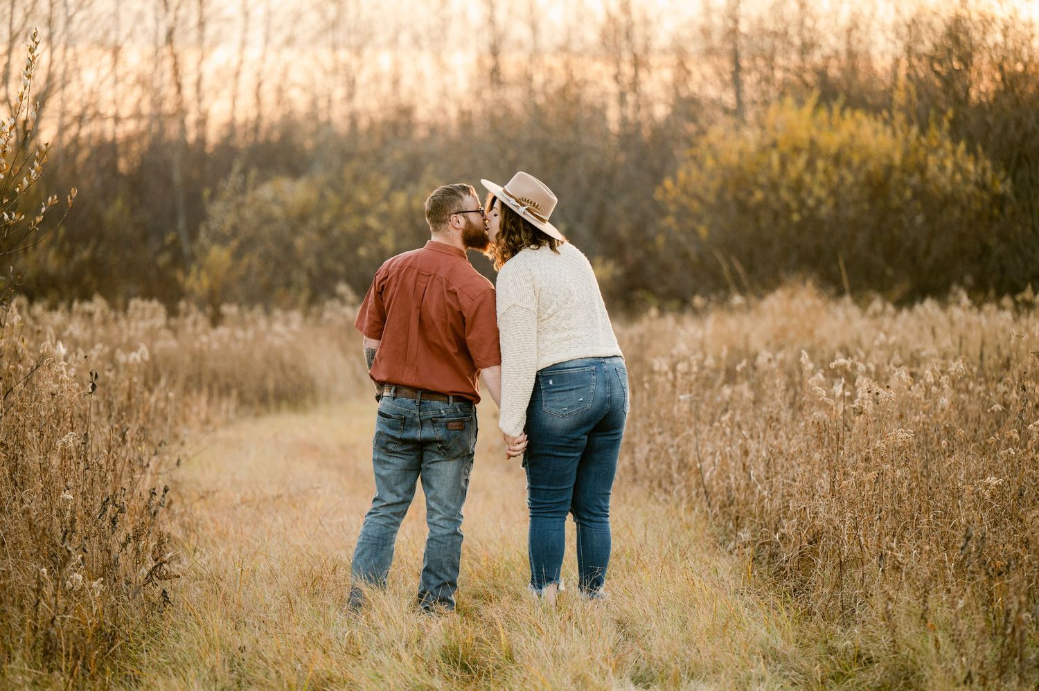 A couple embraces while standing in a golden field at sunset, surrounded by tall grass and autumn foliage.