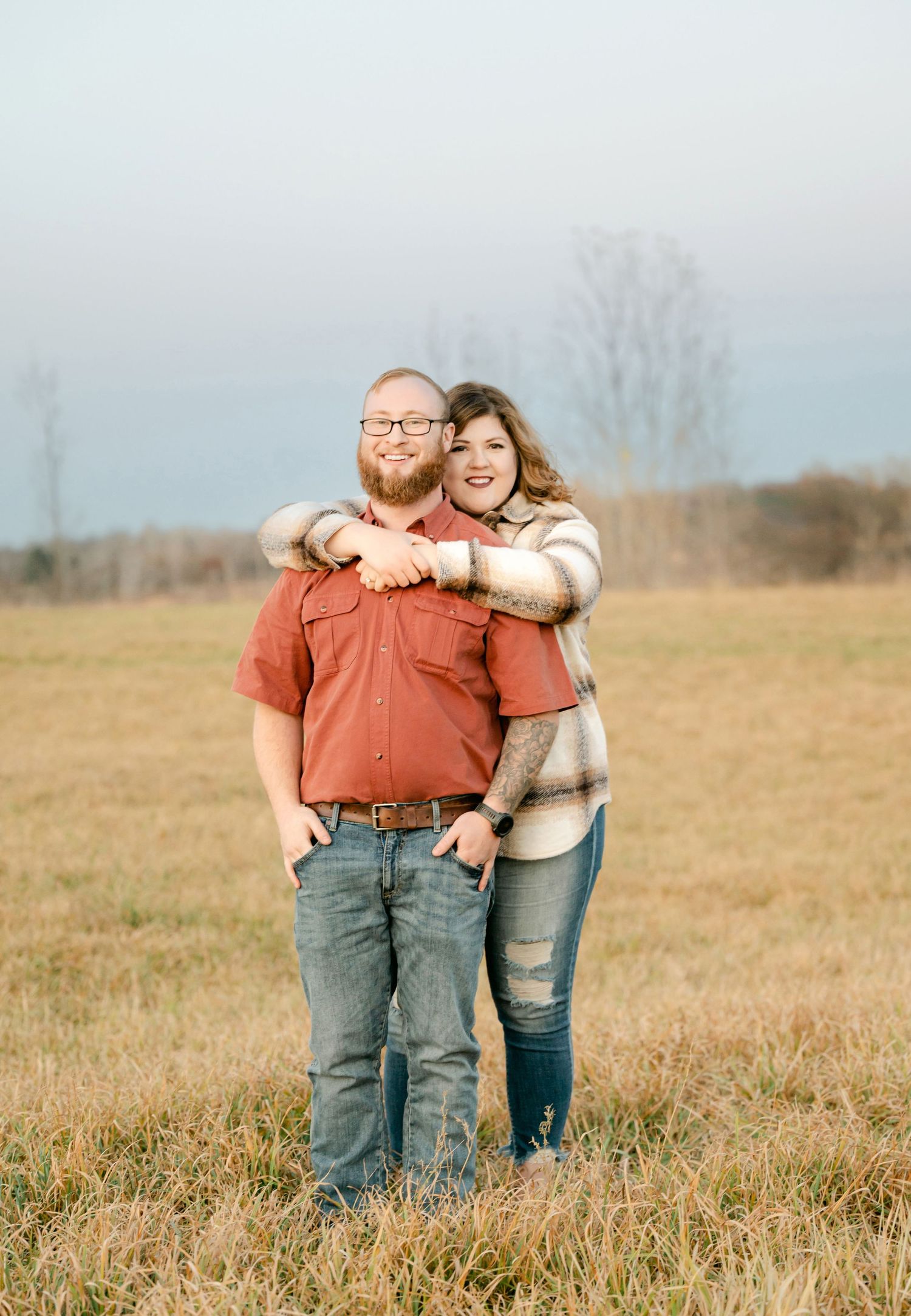 Couple standing together in casual attire in an autumn field with bare trees in the misty background.