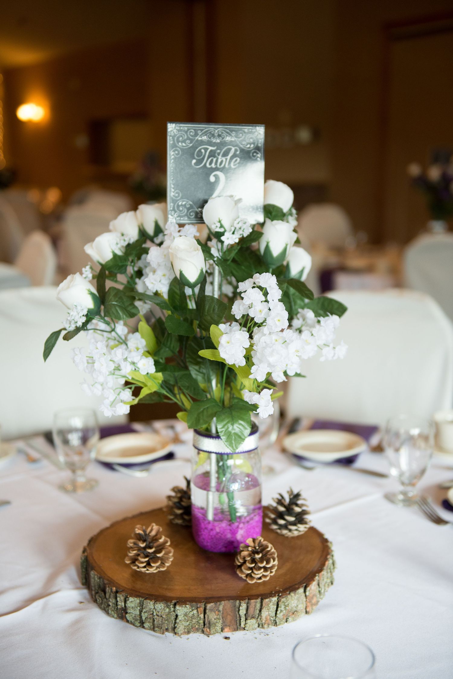 Wedding centerpiece with white flowers and greenery in a purple-dipped mason jar on a rustic wood slice with pinecones. Andover wedding at M