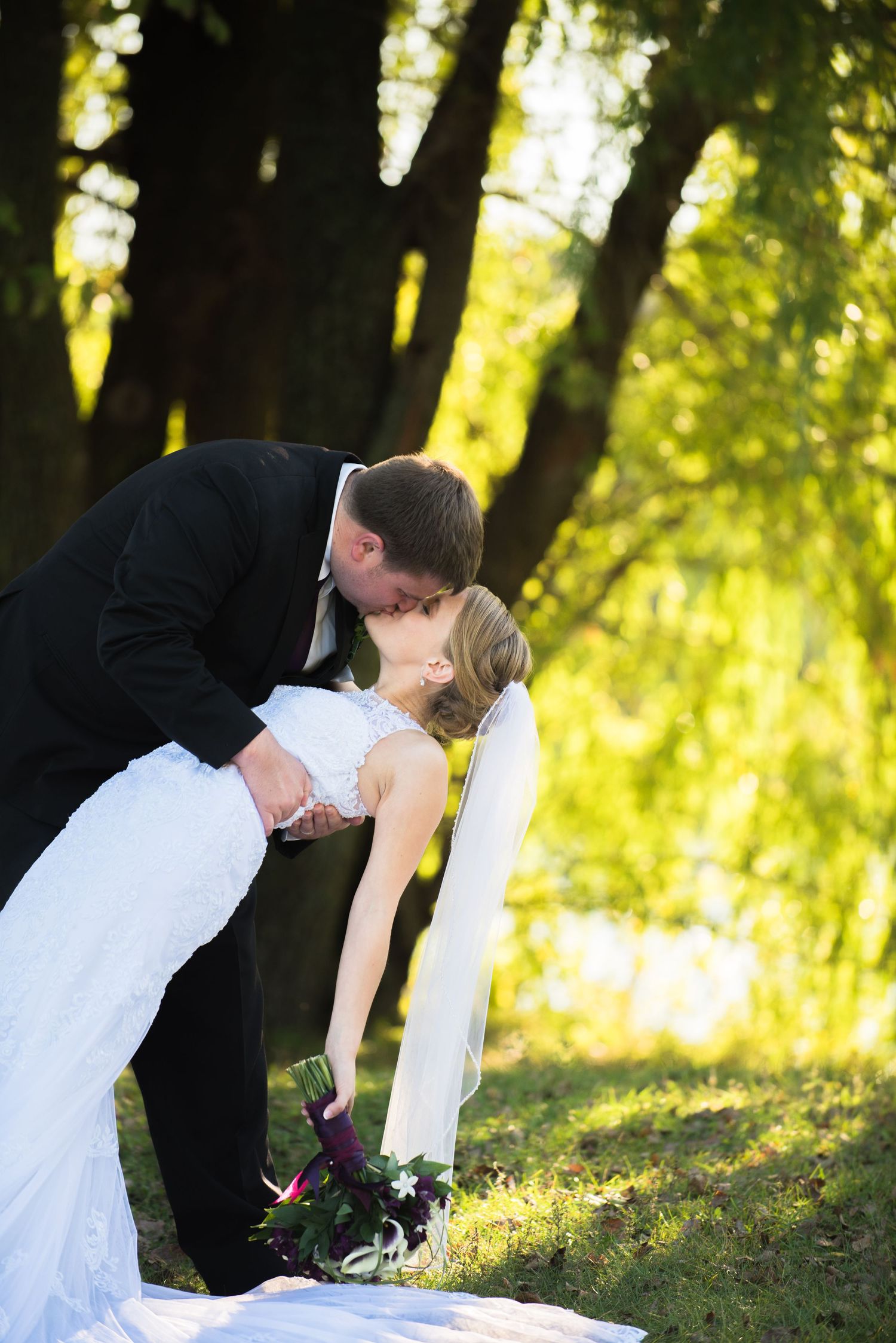 Dramatic dip kiss between newlyweds in a sunlit forest setting. Andover wedding at Magestic Oaks Golf Club