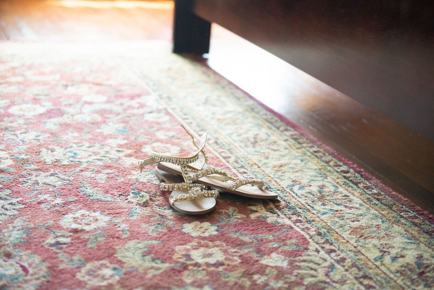 Ballet slippers rest on an ornate red patterned carpet next to dark furniture in soft natural lighting.