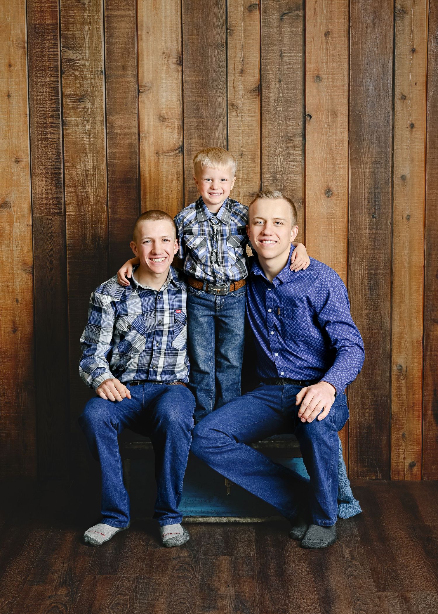 Family group in coordinating blue outfits sitting against a rustic wooden wall backdrop.
