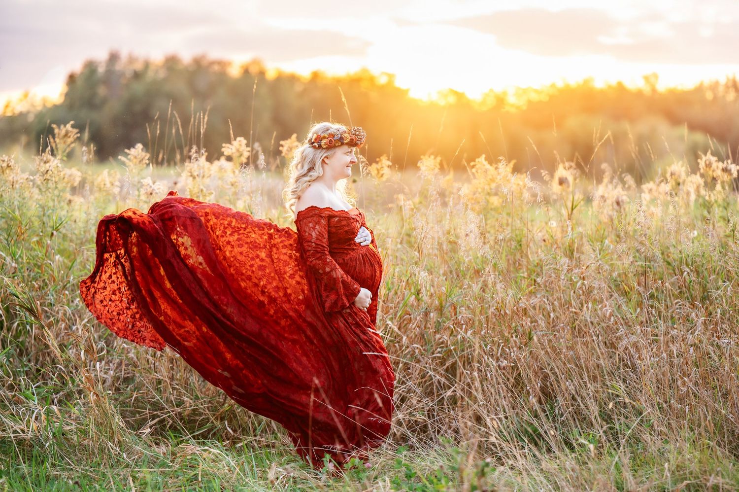 A pregnant woman in a flowing red dress twirls in a golden field at sunset.