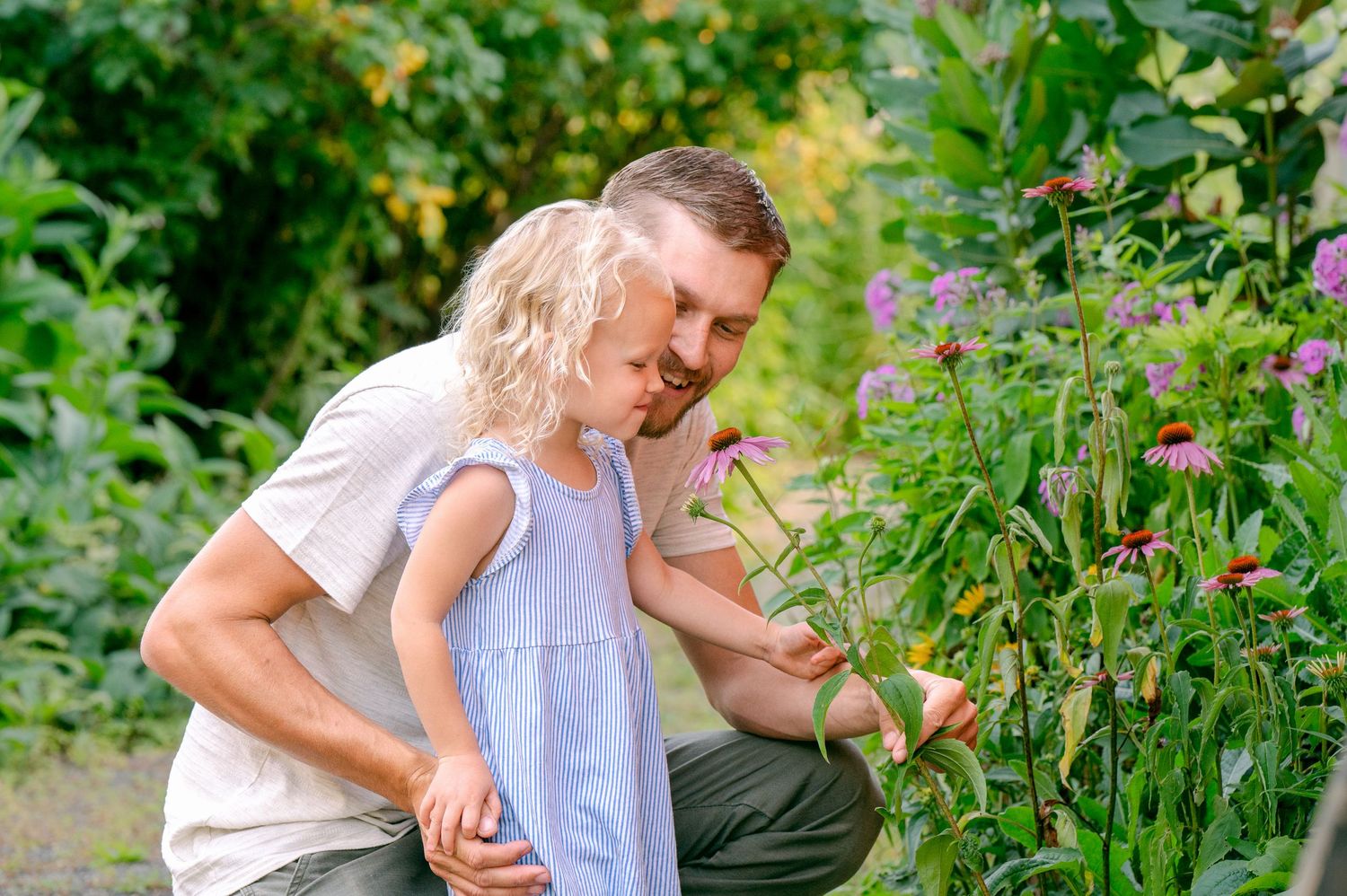 Two figures share a tender moment while examining flowers in a verdant garden setting.