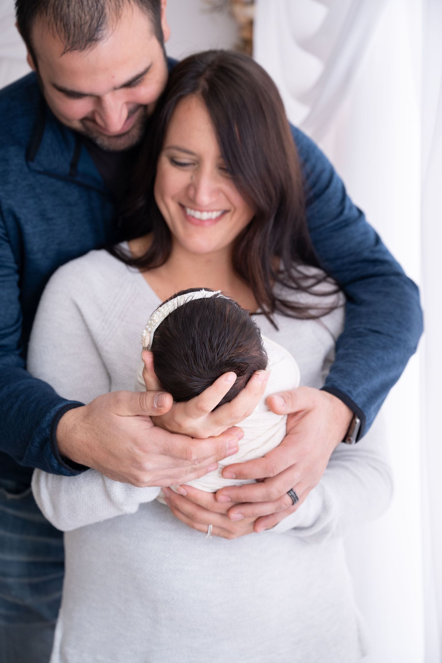 Happy parents embracing while holding their newborn baby in white room.