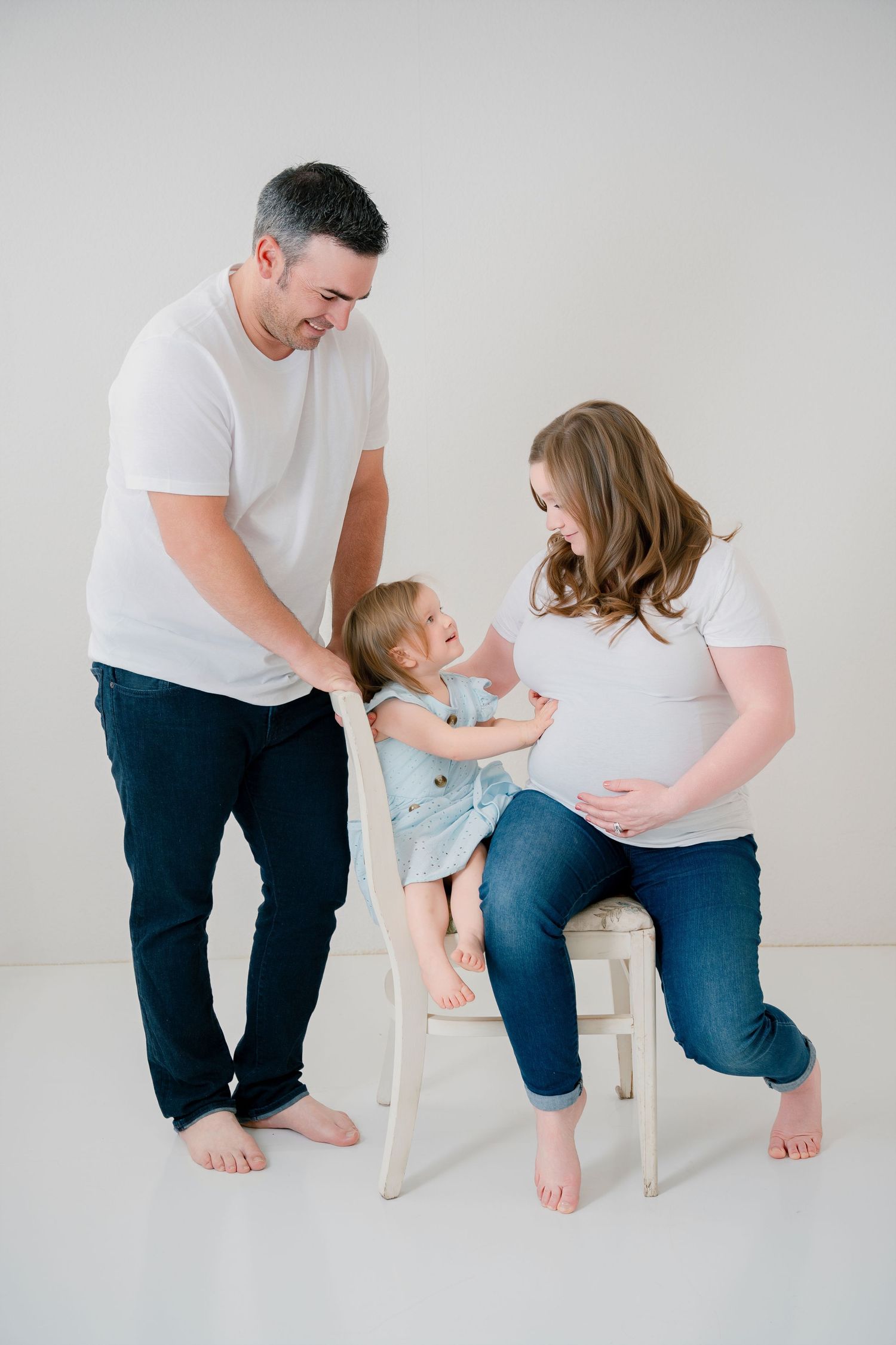 Family gathers around white chair for indoor maternity photo with toddler in light blue dress.
