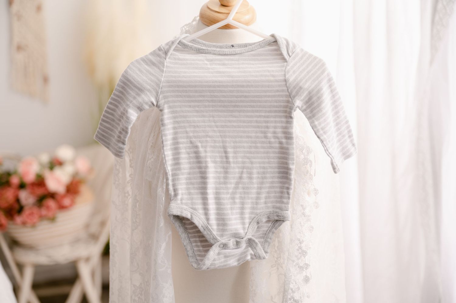 A gray and white striped baby onesie hanging by a window with soft natural light and pink flowers in the background.