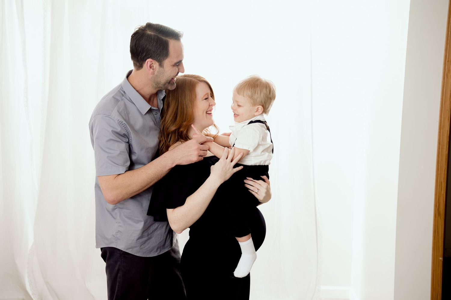 A couple wearing casual clothes share a tender moment with their toddler child against a white curtain backdrop.