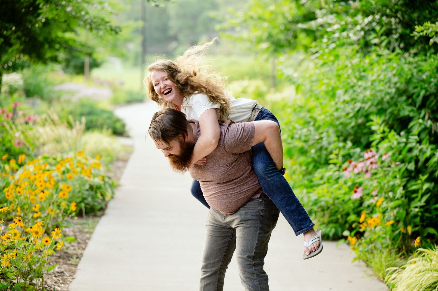 Couple enjoying a carefree moment with piggyback ride on pathway surrounded by colorful summer flowers.