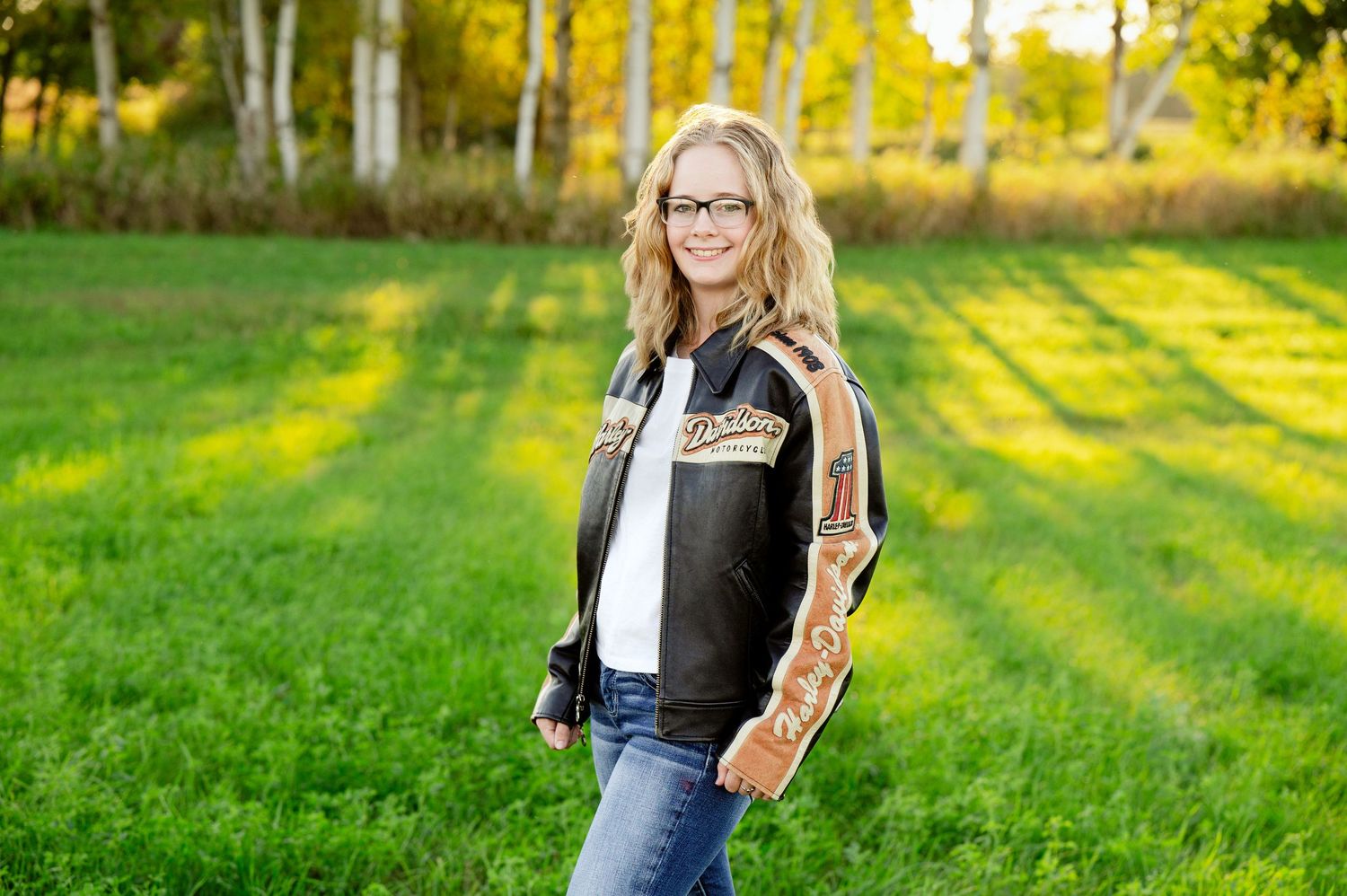 Senior Portrait in Foreston MN Person posed in casual denim outfit against backdrop of sunlit grass and trees.
