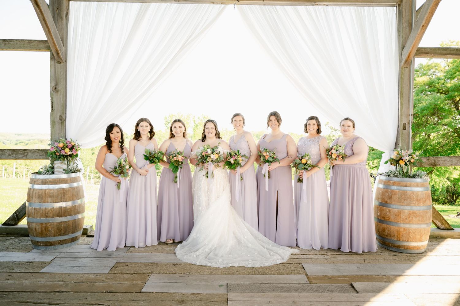 Wedding party poses under white draped pavilion with rustic wine barrels and lilac bridesmaid dresses.