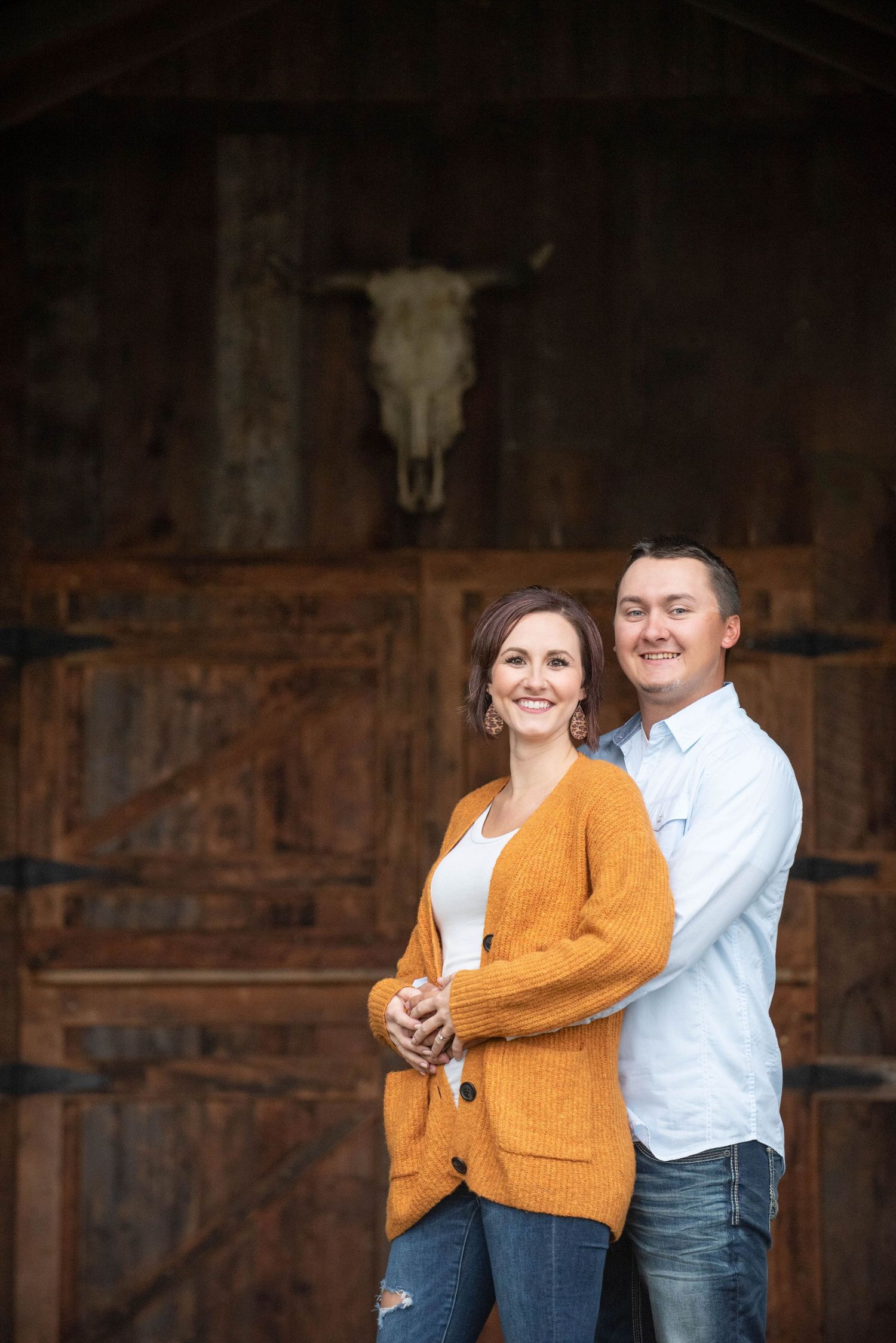 Romantic rustic barn engagement photos show couple wearing white shirt and mustard cardigan embracing against wooden backdrop.