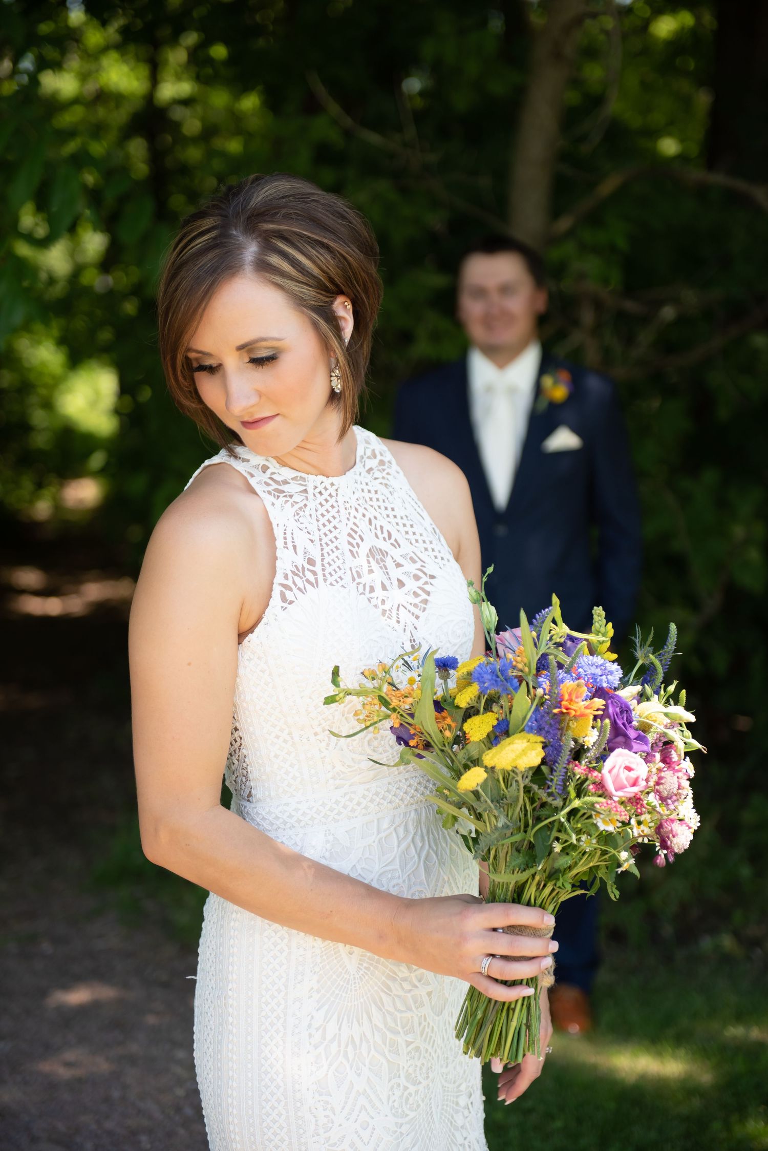 Bride in a white lace dress admires her colorful wildflower bouquet in a sunlit outdoor garden setting.