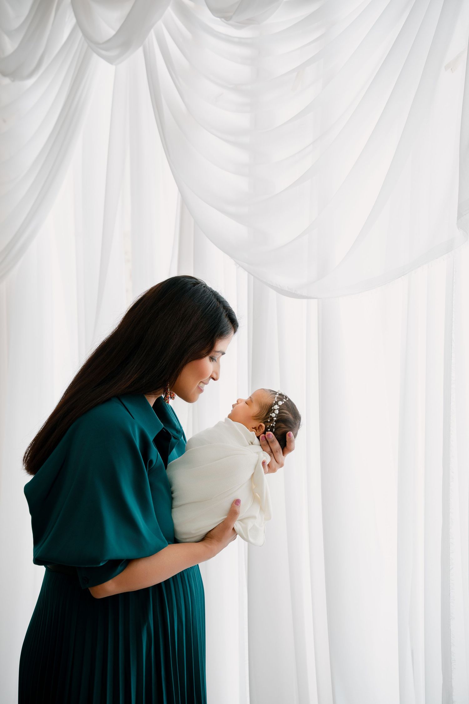 Woman in teal dress holds baby against white draped backdrop during photo session.