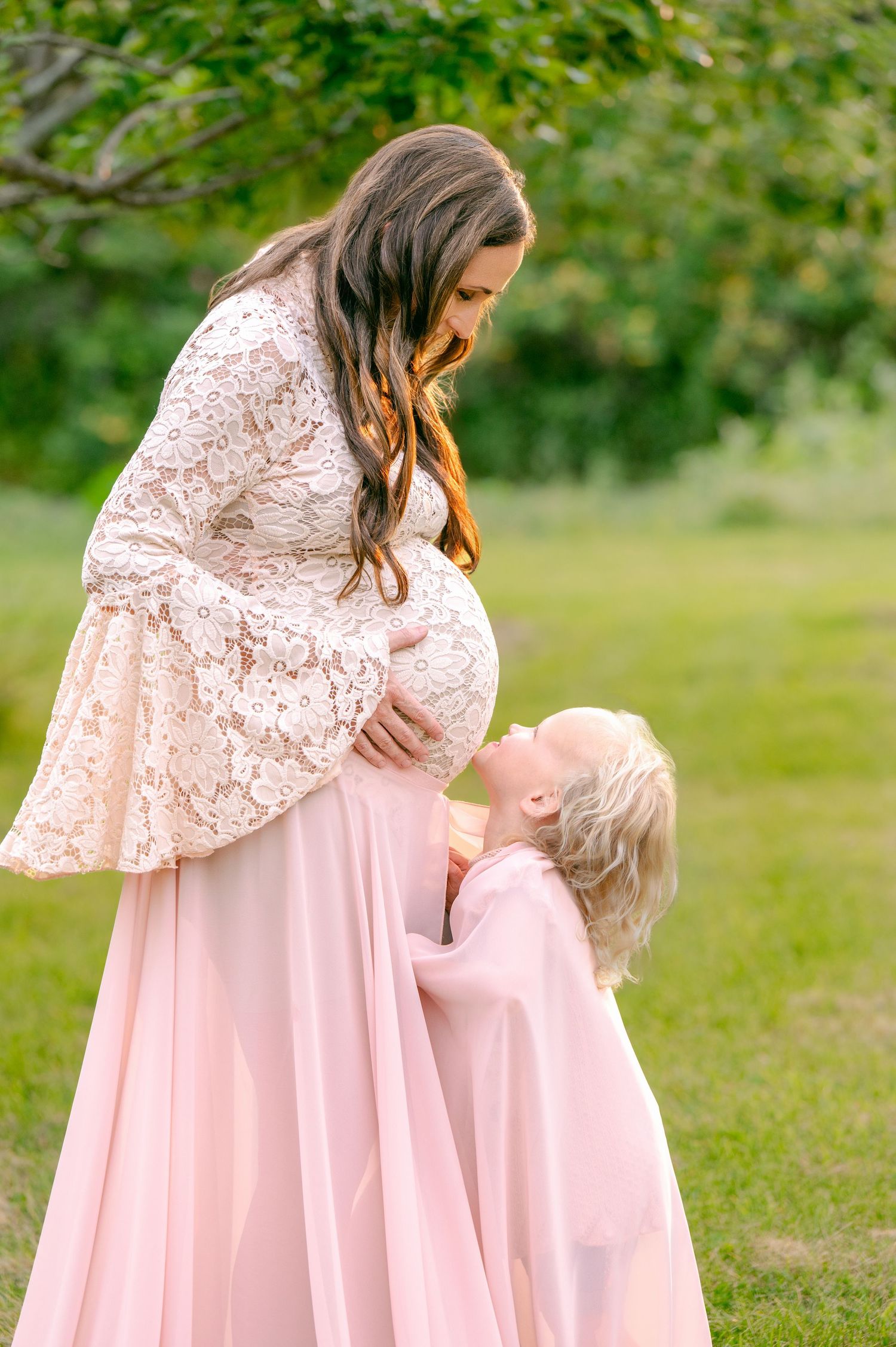A pregnant woman in a pink lace dress sharing a tender moment with a small child in a matching outfit outdoors.