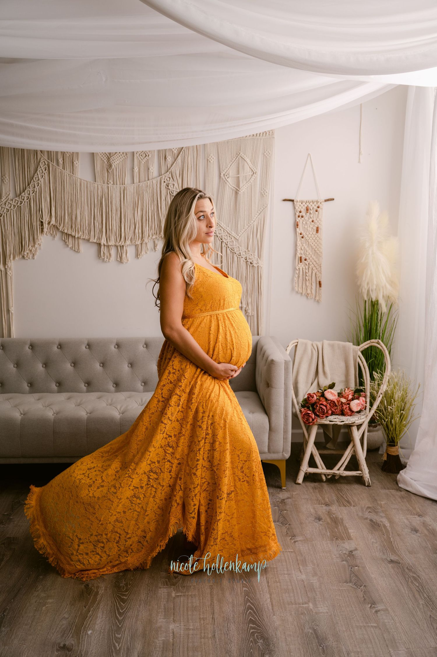 A pregnant woman in a flowing mustard yellow lace dress poses in a bohemian styled room with macrame wall decor.