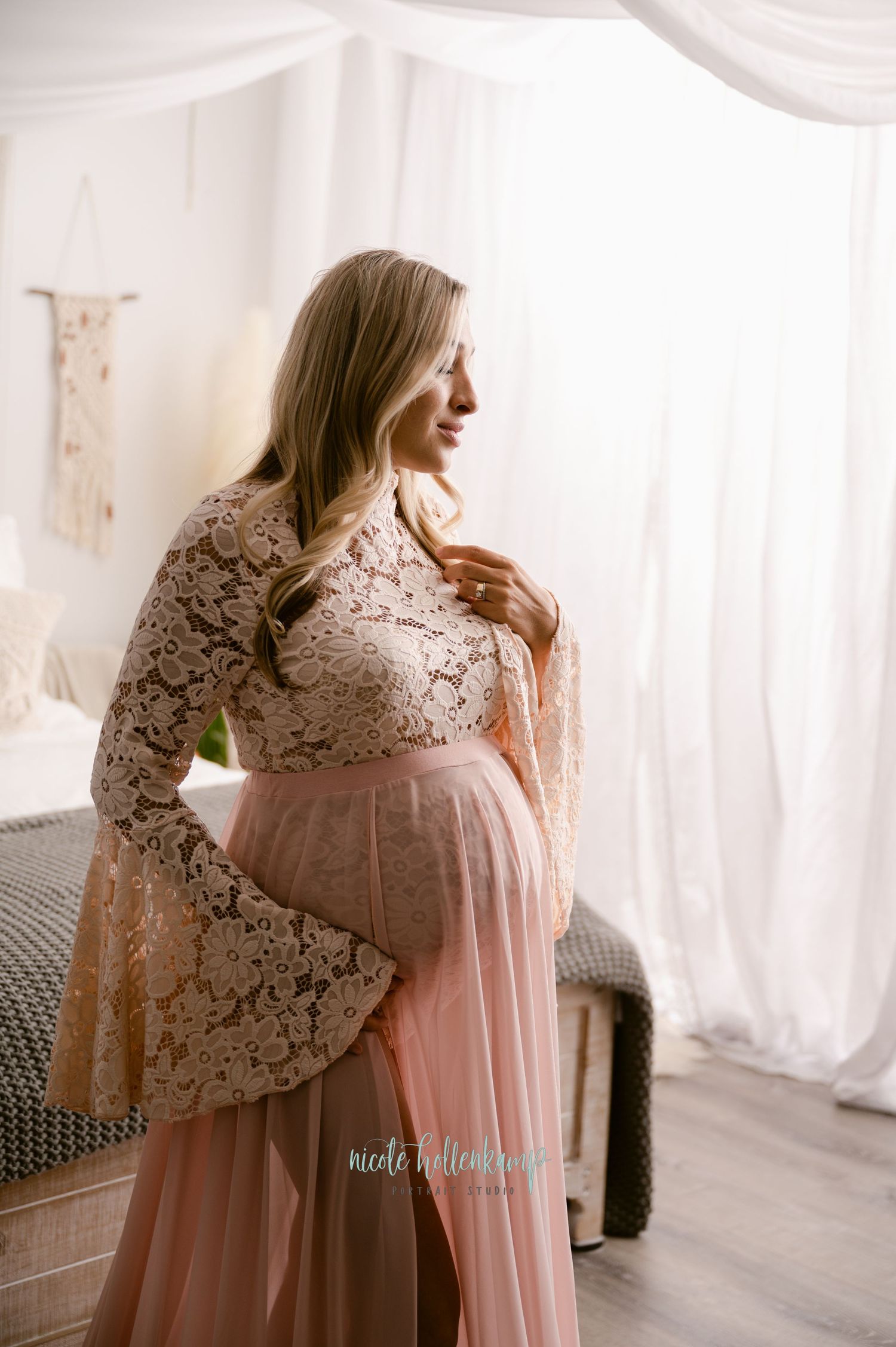 Profile view of an expectant mother in blush pink gown with white lace bell sleeves in a bright studio setting.