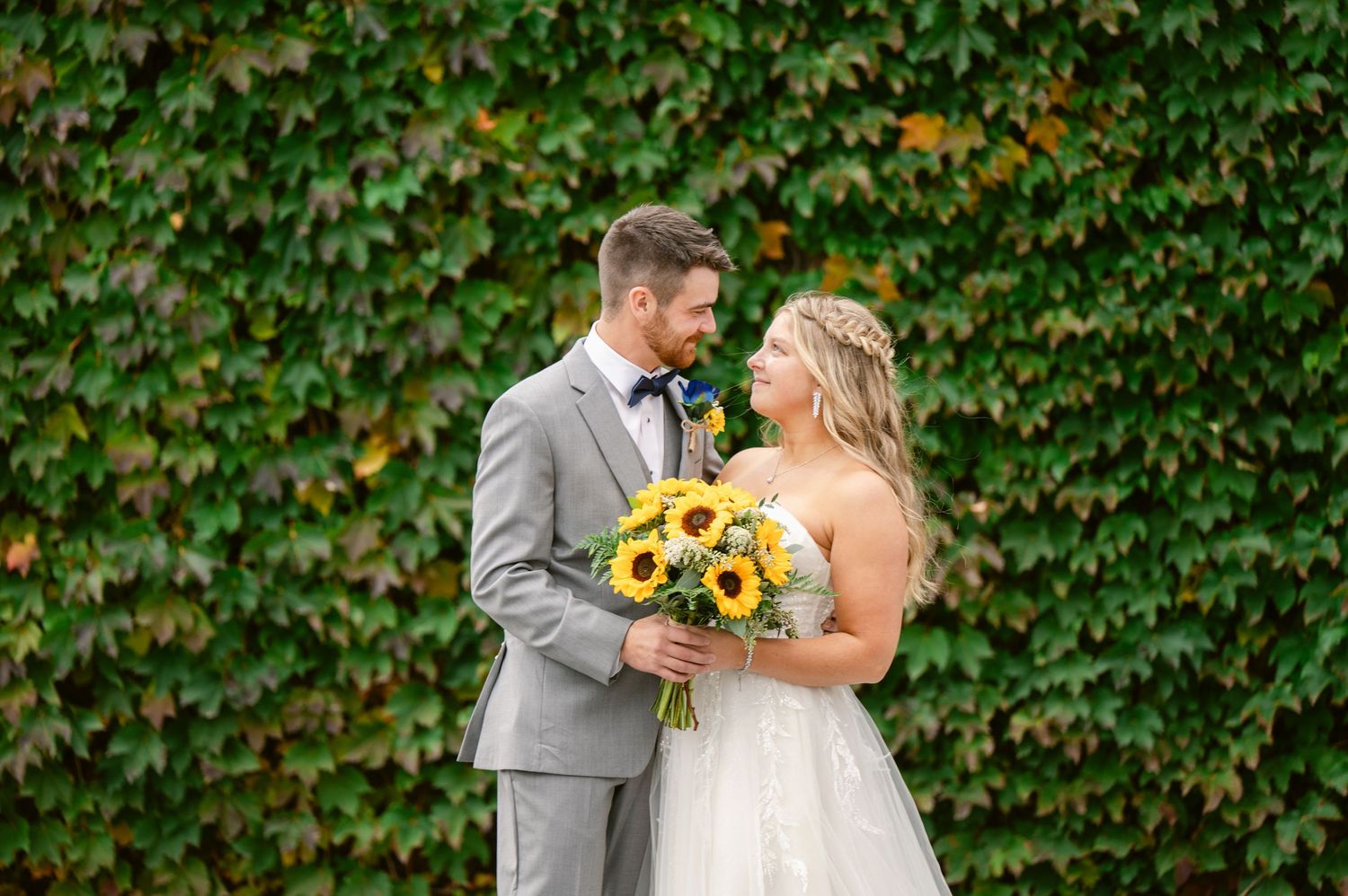 A couple in wedding attire share a romantic moment against an ivy-covered wall, with the bride holding bright sunflowers.