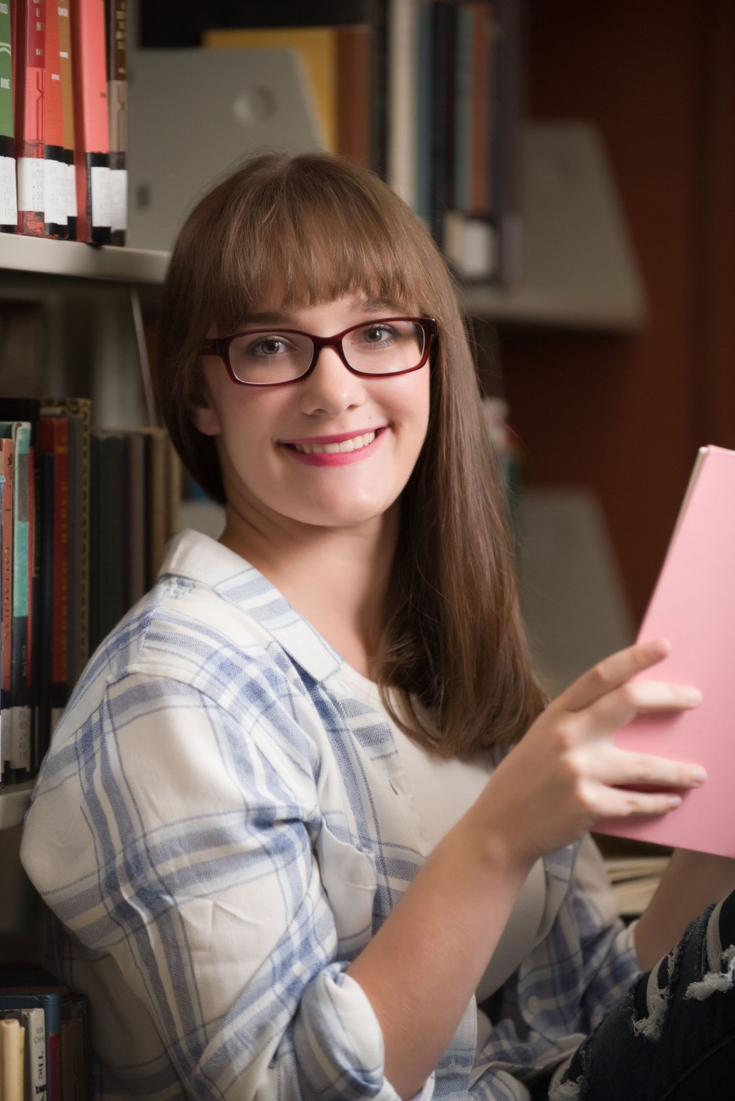 Senior Portrait at St Cloud State College in glasses and plaid shirt smiling while holding pink paper in library setting.
