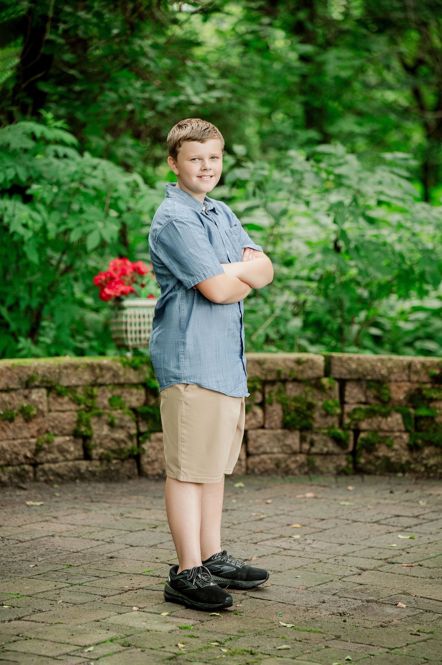 Young child in a blue shirt and khaki shorts standing on brick pavement near garden foliage.