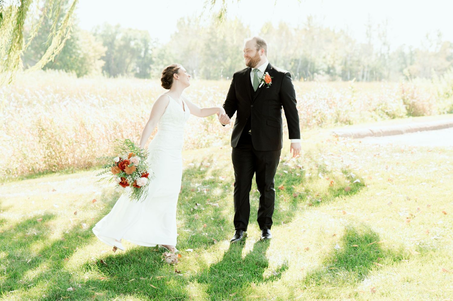The Grands At Mulligans. A wedding couple takes a romantic stroll in a grassy meadow on a bright, sunlit day.