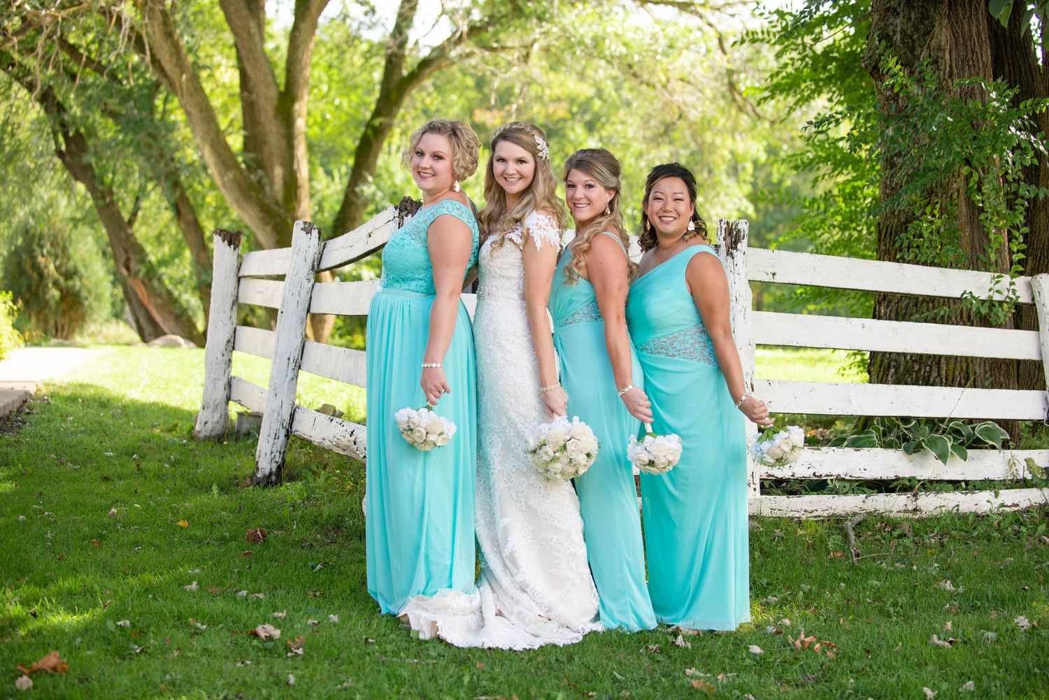 A bridal party in matching turquoise gowns poses outdoors against a white fence. Rum River Barn Milaca Minnesota Weddings