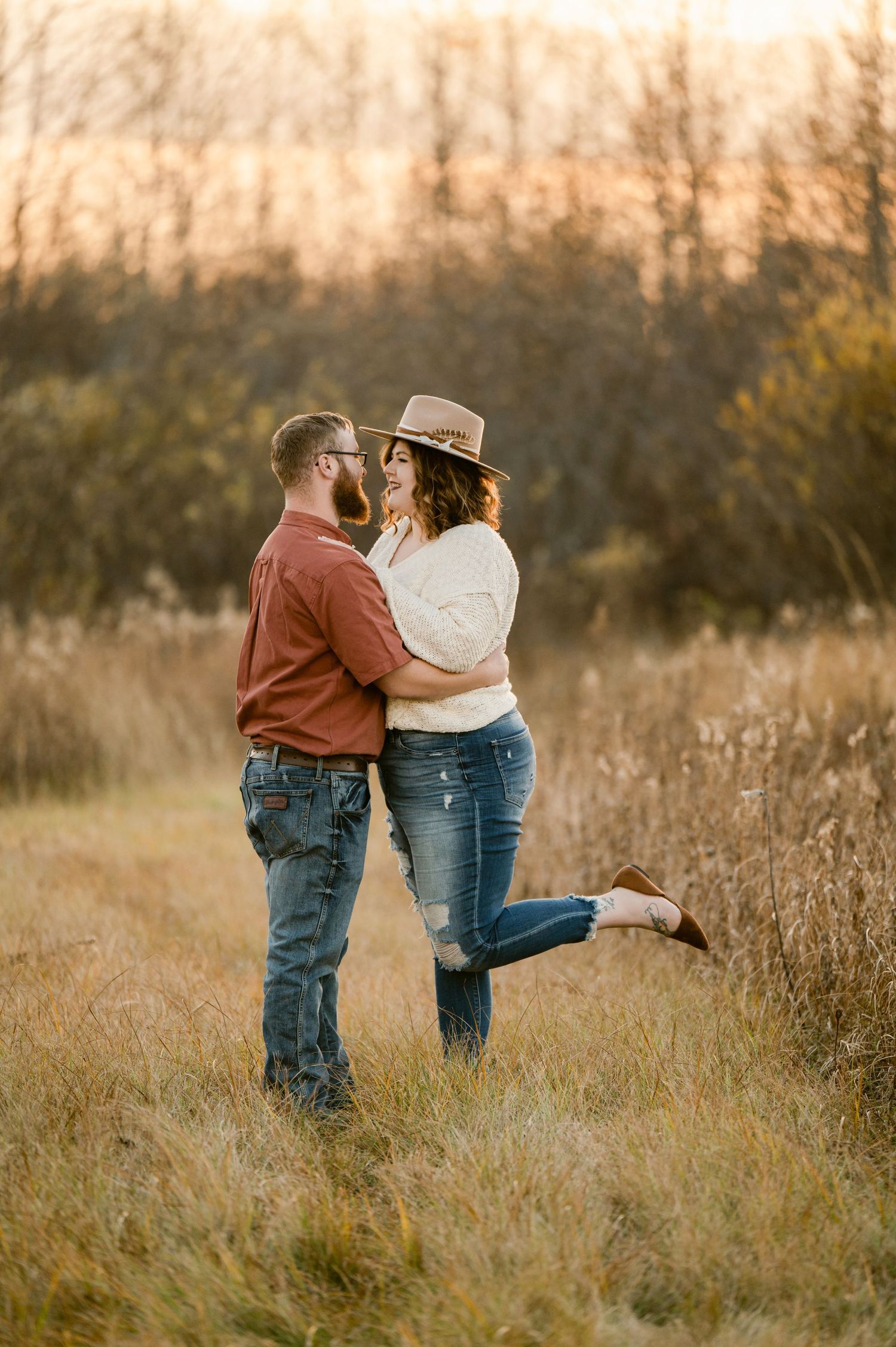 A romantic couple embraces in a golden field at sunset wearing rustic casual attire and a brown hat. St cloud area engagement photos.