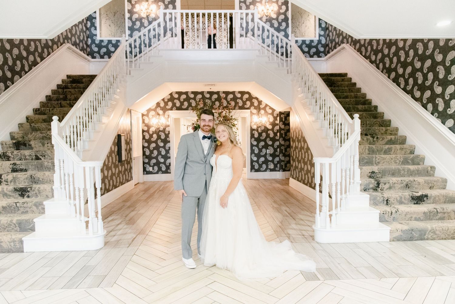 Elegant couple stands beneath grand double staircase with ornate white railings and patterned wallpaper in venue ballroom.