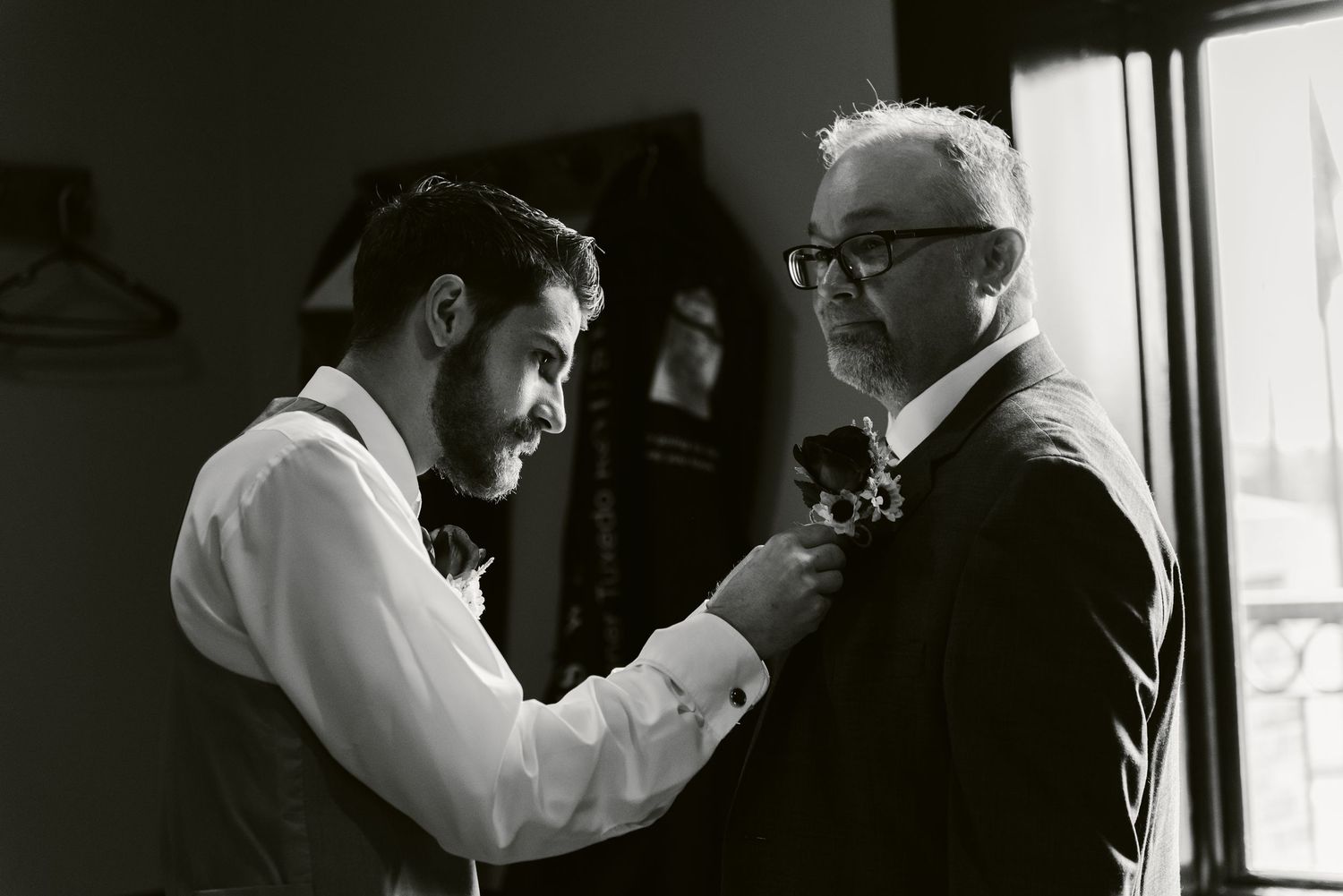 A black and white photo shows someone helping adjust another person's bow tie in a dimly lit room.