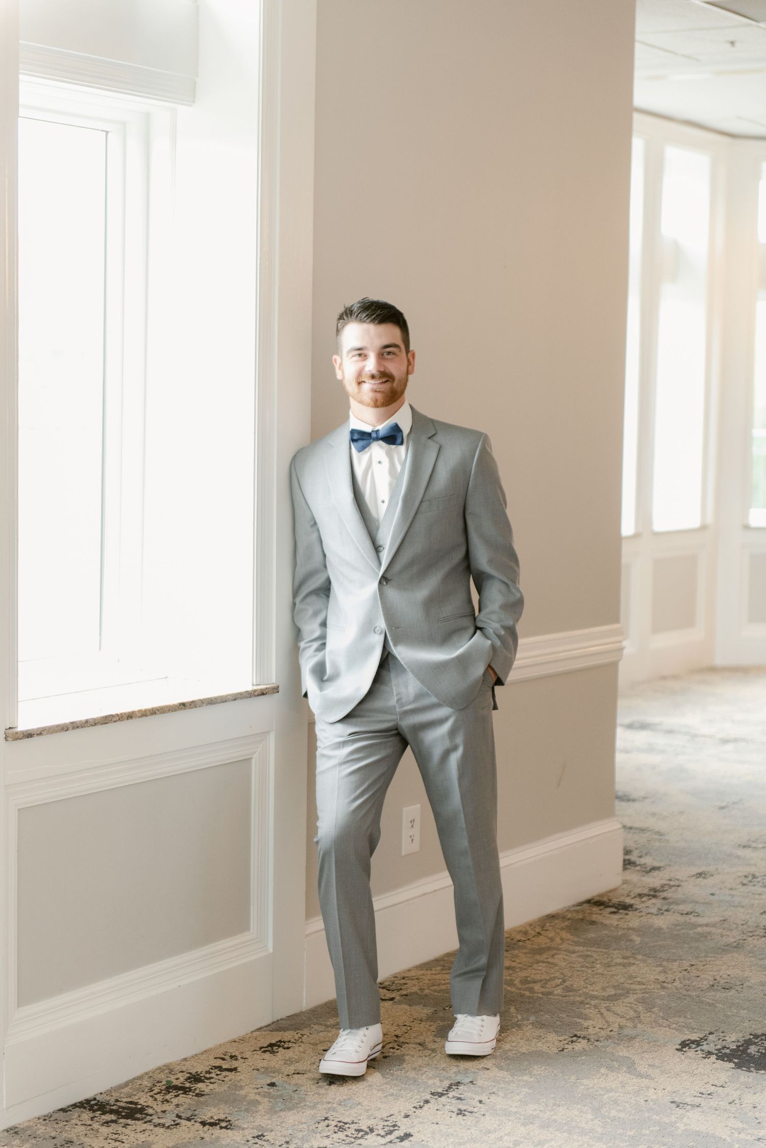 A person in a light grey suit and blue bow tie stands confidently against a beige wall.
