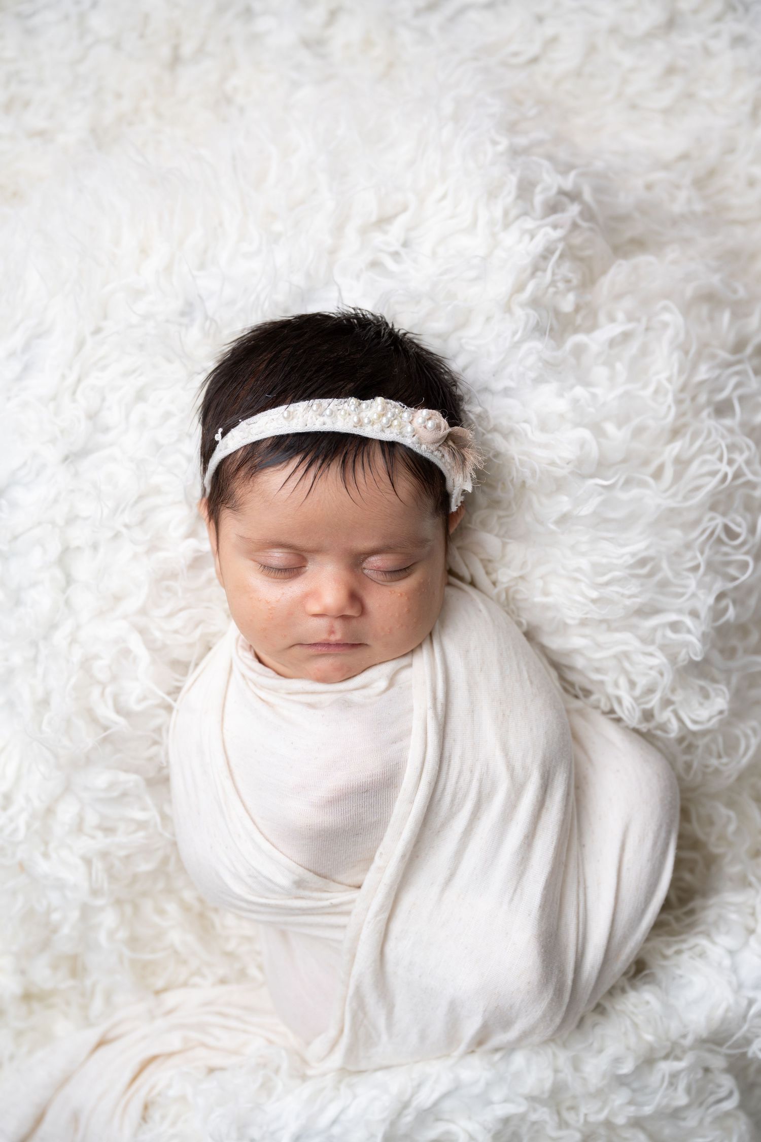 Sleeping newborn swaddled in white with lace headband on fluffy white blanket.