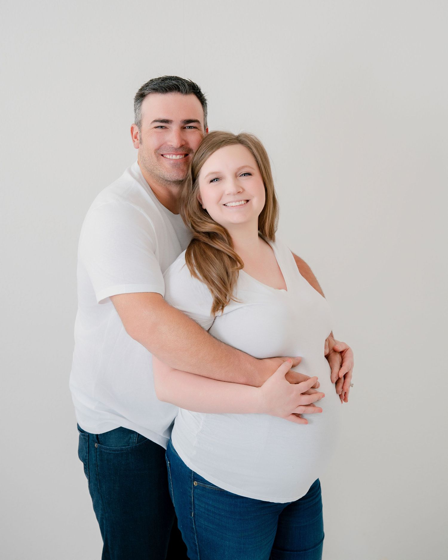 Couple embraces in studio setting wearing white shirts for maternity photo.