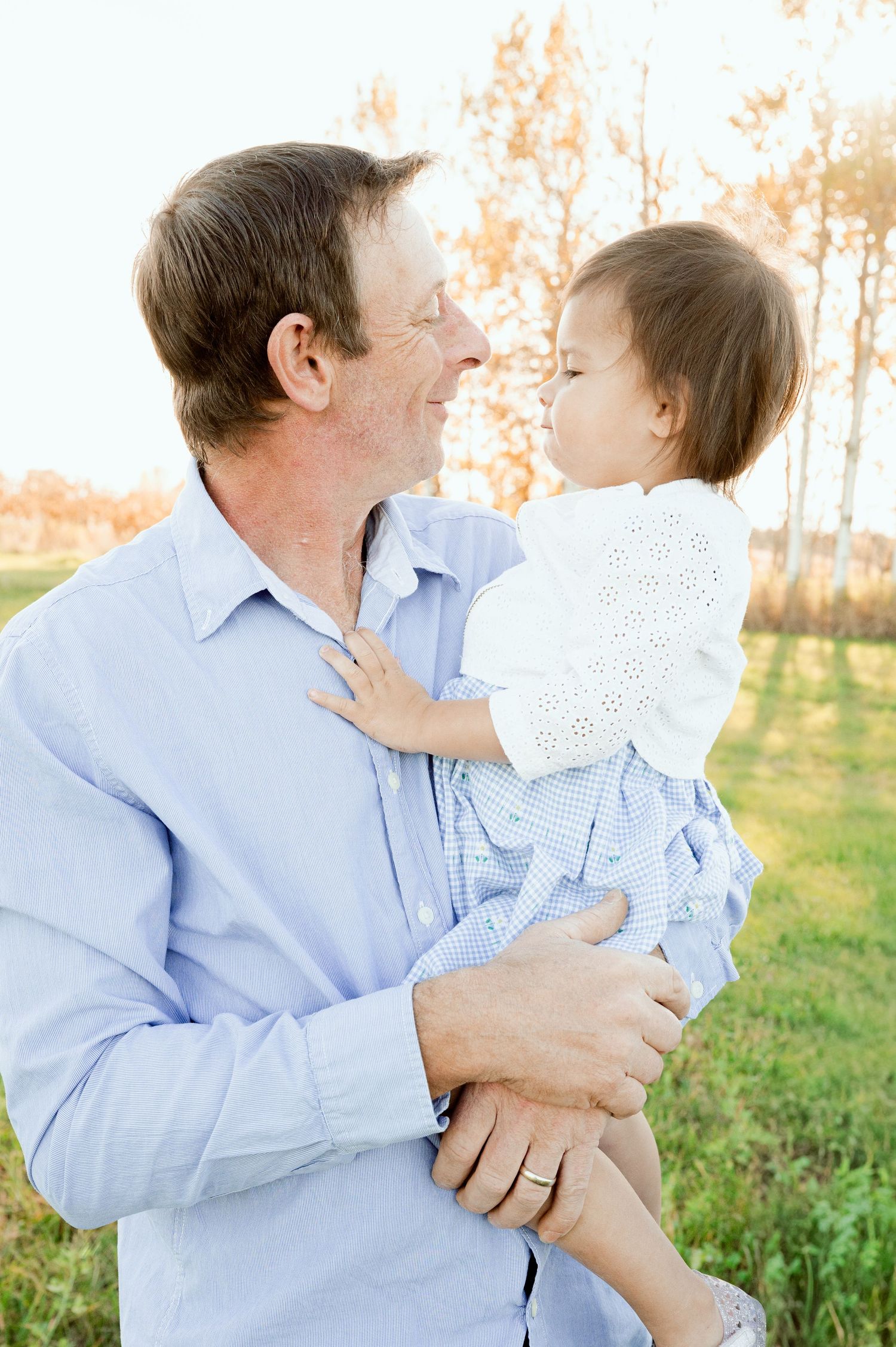 A parent holds a young child wearing eyelet lace against a natural outdoor backdrop.