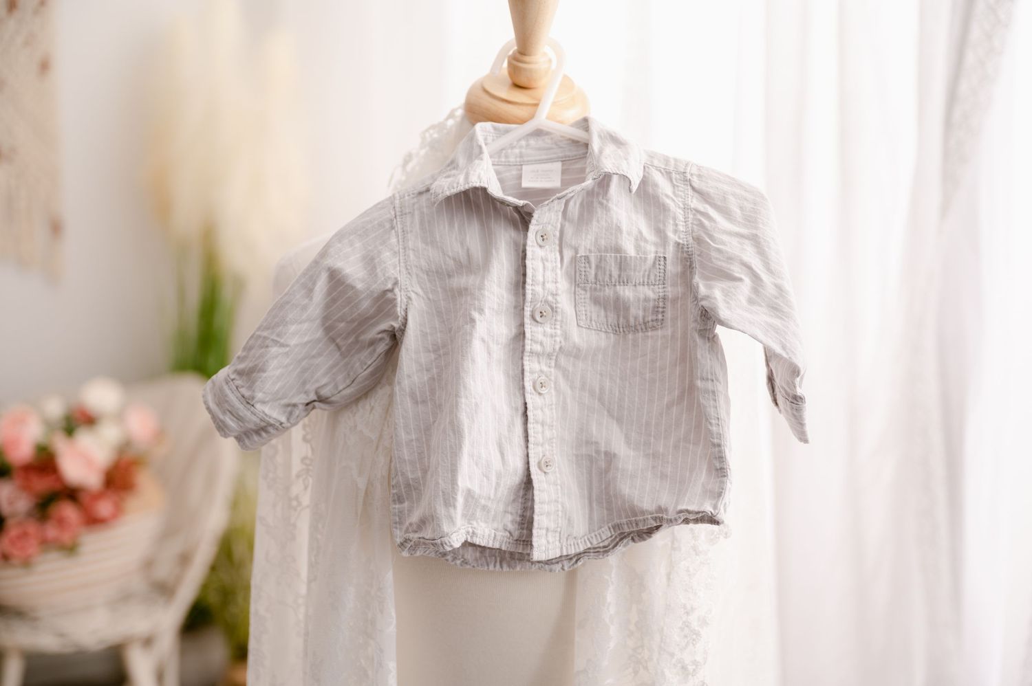 Small striped gray and white button-down baby shirt hanging on wooden hanger against sheer white curtain background.