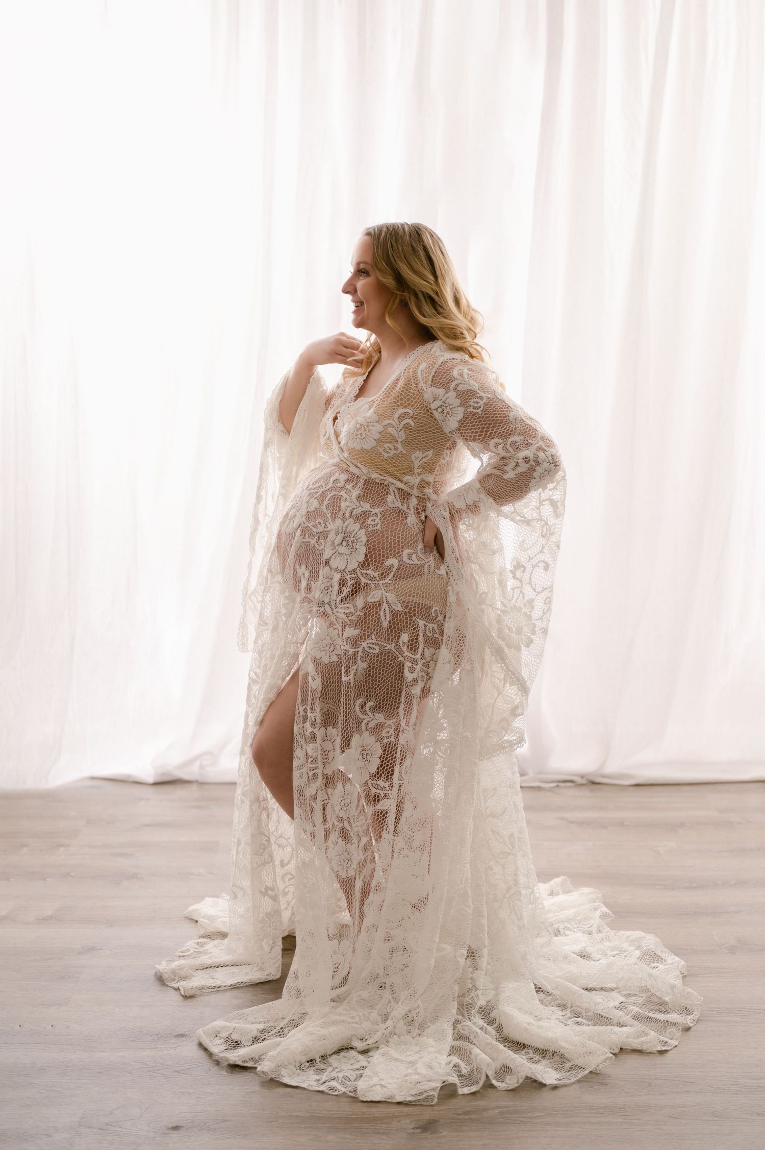 Elegant maternity gown in white lace with long flowing sleeves and train photographed against light curtain backdrop.