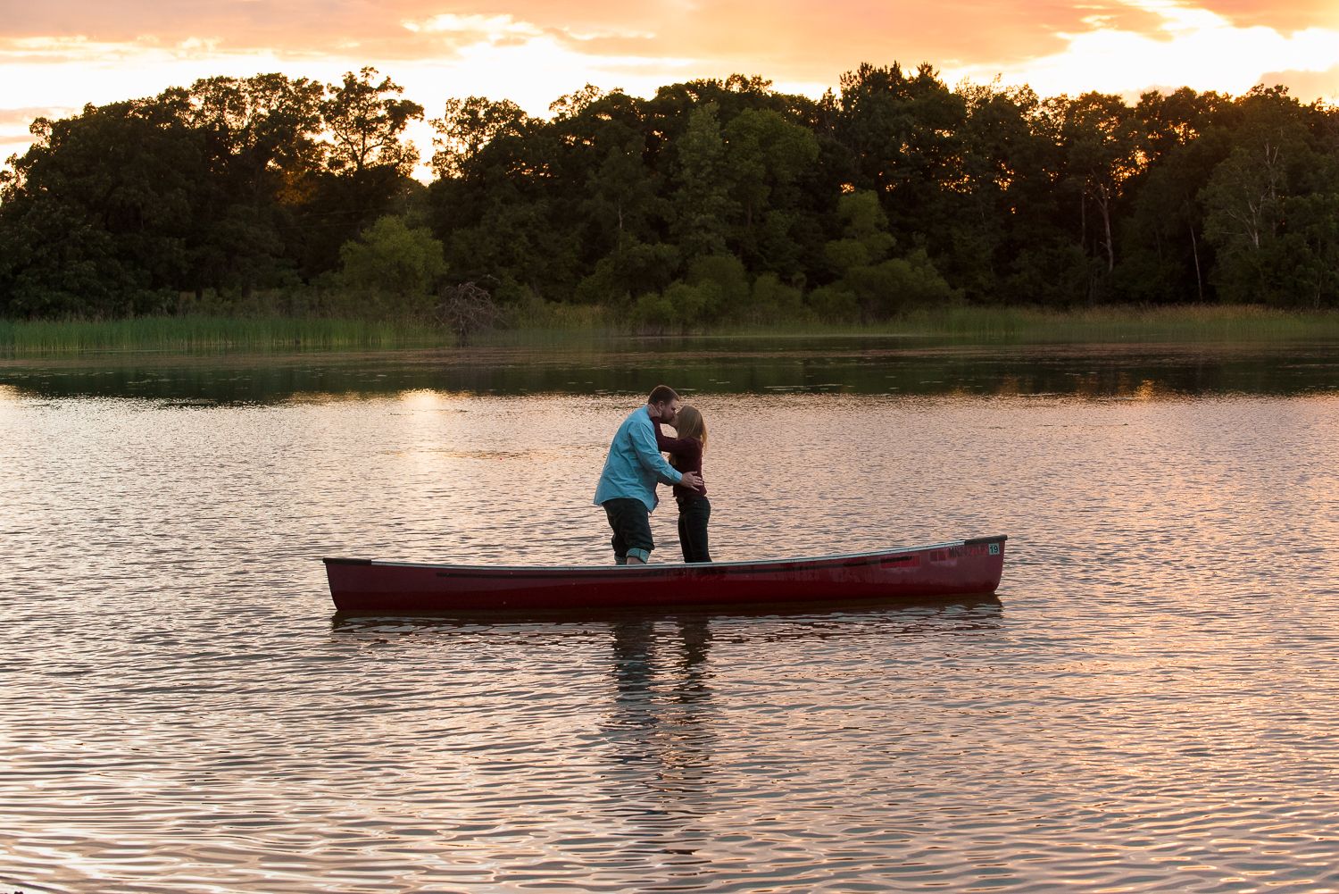 Silhouettes of two people in a red canoe gliding across a tranquil lake at sunset creating a romantic scene.