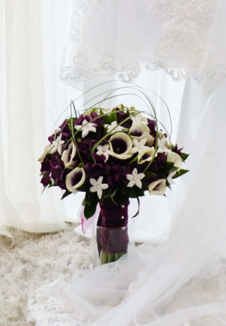 Elegant bridal bouquet with white calla lilies, dark purple flowers and small white stephanotis blooms against lace backdrop. Andover weddin