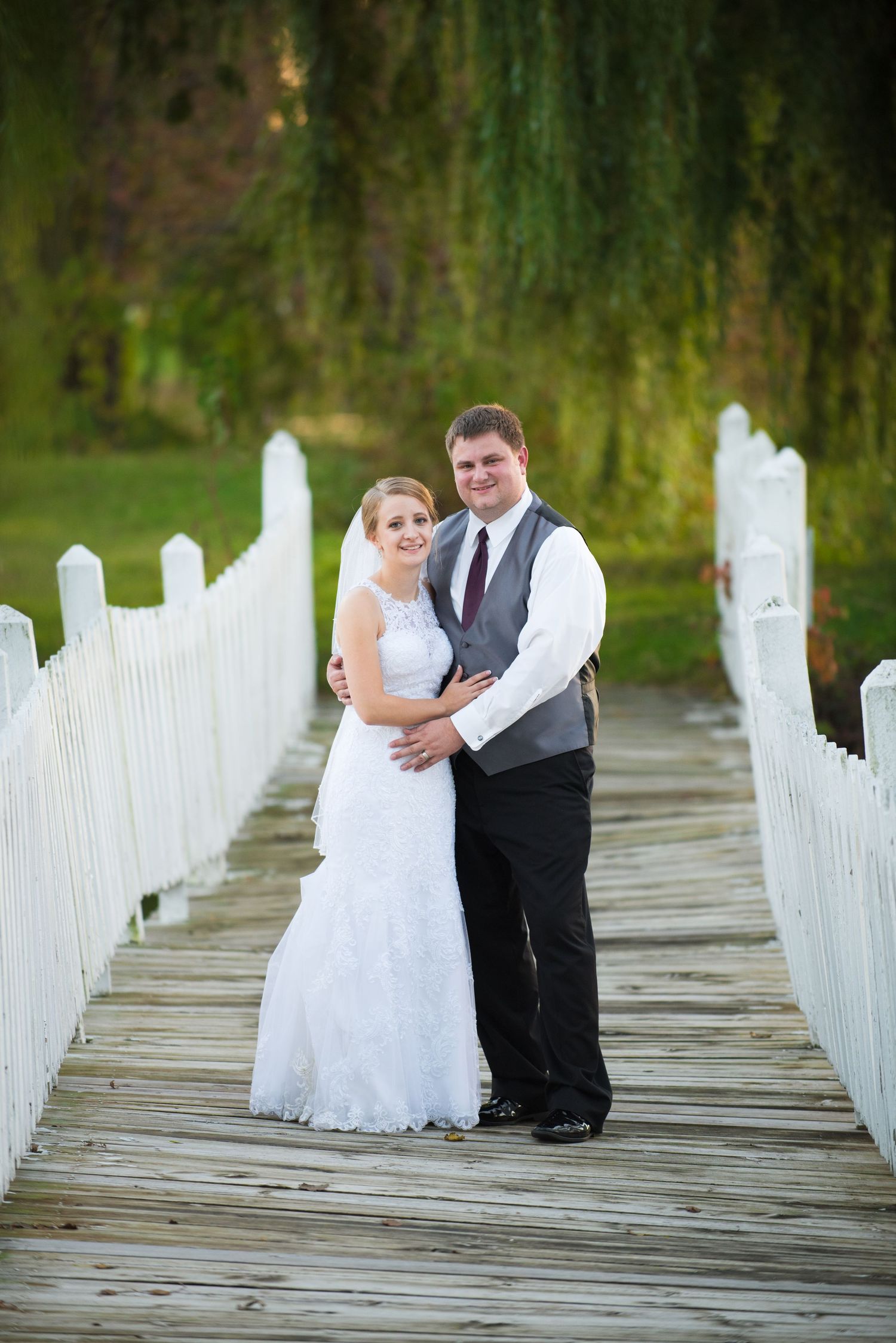 A newlywed couple poses on a wooden bridge with white railings and weeping willows in the background. Andover wedding at Magestic Oaks Golf