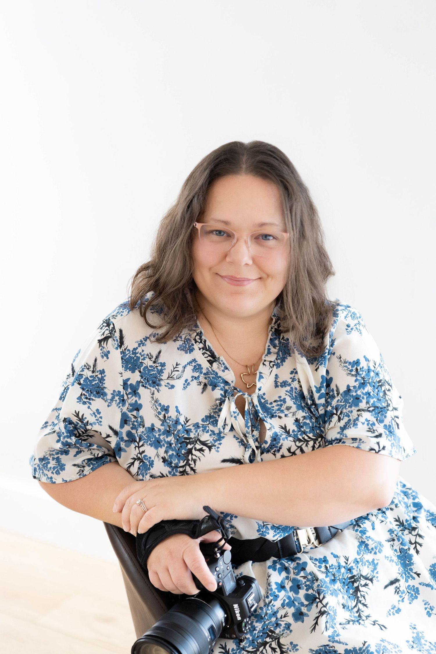 Professional photographer in white and blue floral dress sitting with camera equipment against light background.