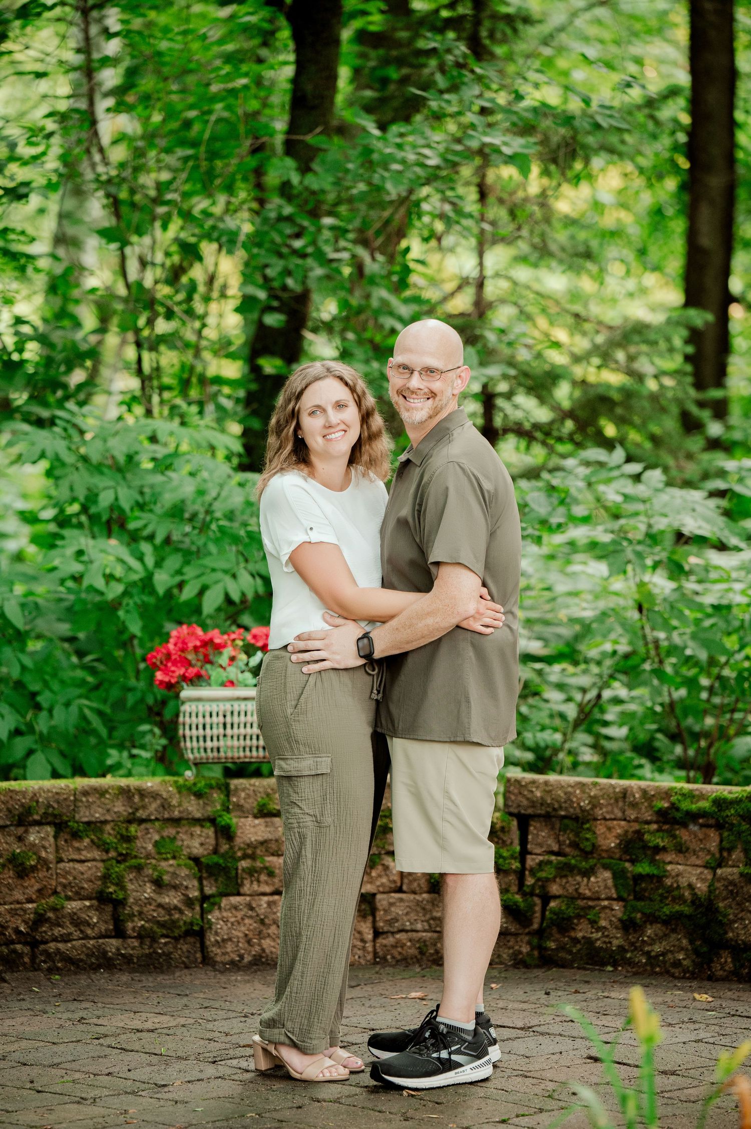 Clear Lake MN Family Photos: A loving couple poses for a portrait on a stone patio with red flowers and green foliage in the background.