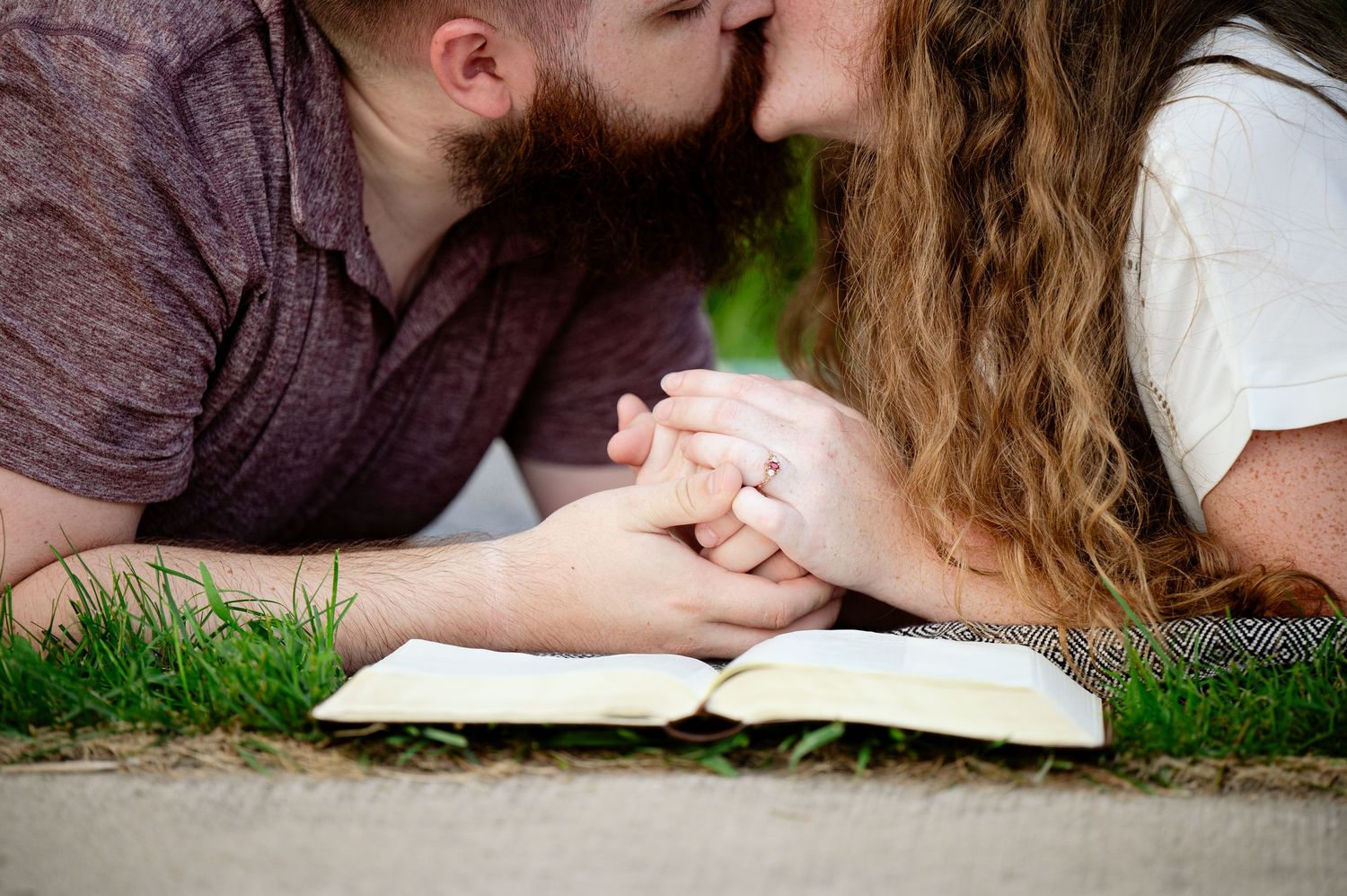 A couple lying in grass holds hands while reading a book together on a sunny day.