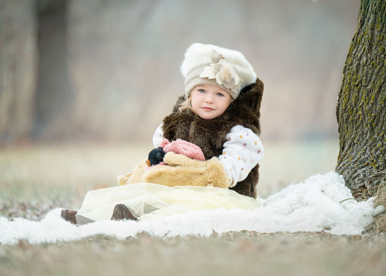 A child sits outdoors in the snow wearing a warm winter coat and crocheted beanie hat near a tree.