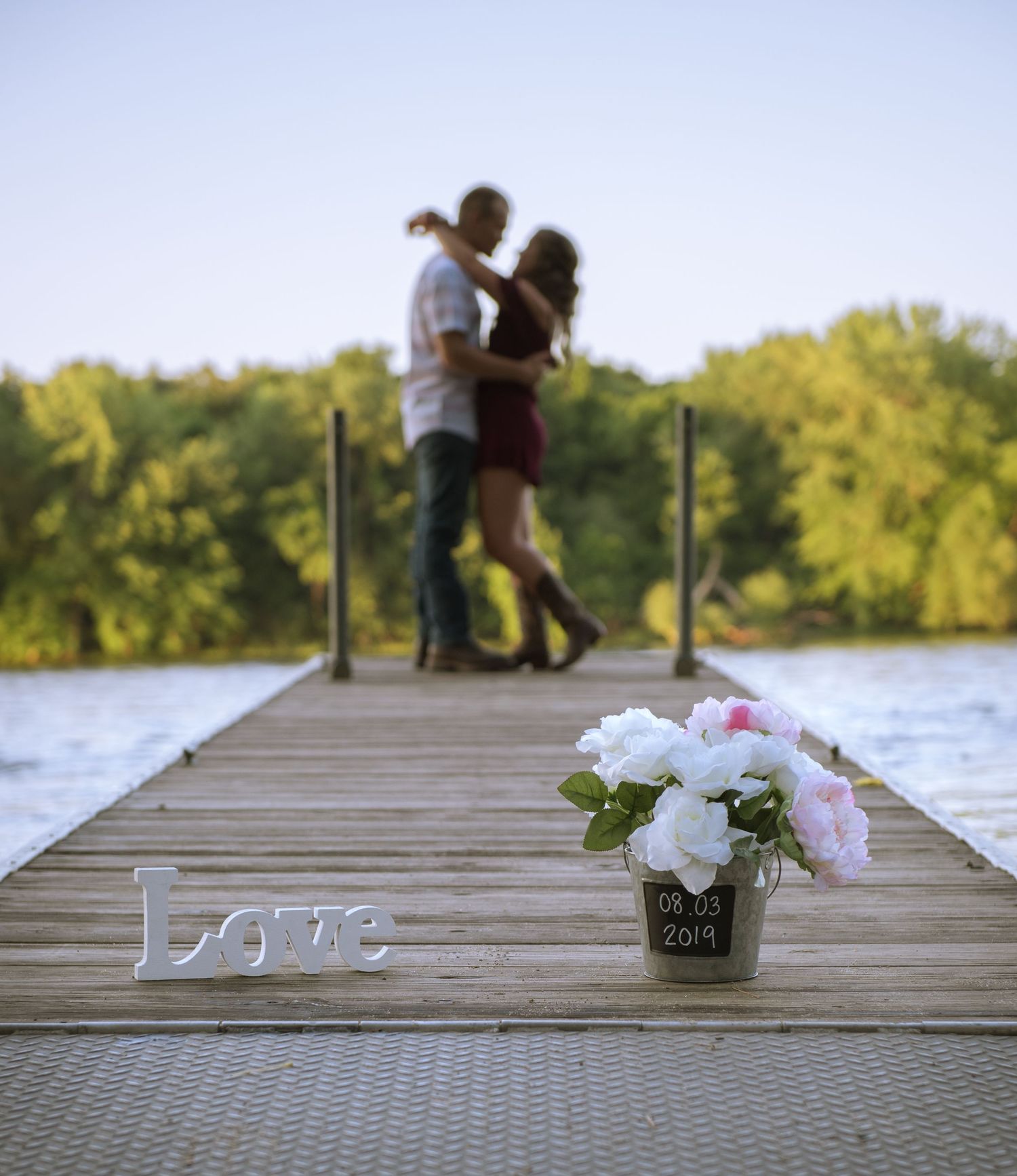 A romantic scene on a dock with a couple embracing in the distance, while 'love' decor and flowers sit in the foreground.