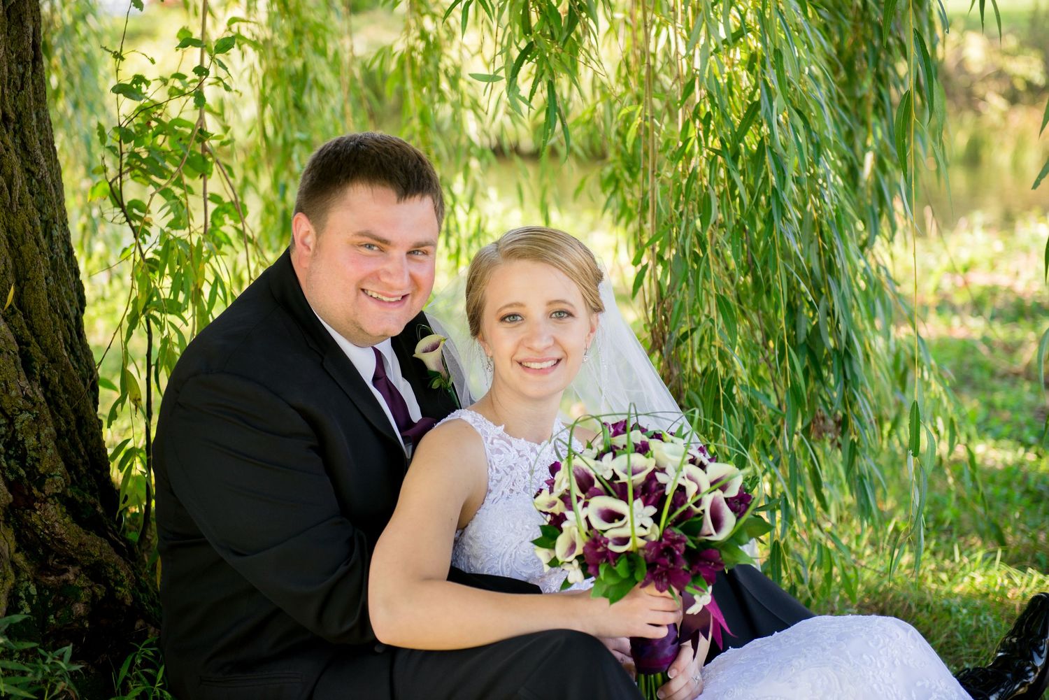 Wedding couple poses together under hanging willow branches with a purple floral bouquet. Andover wedding at Magestic Oaks Golf Club
