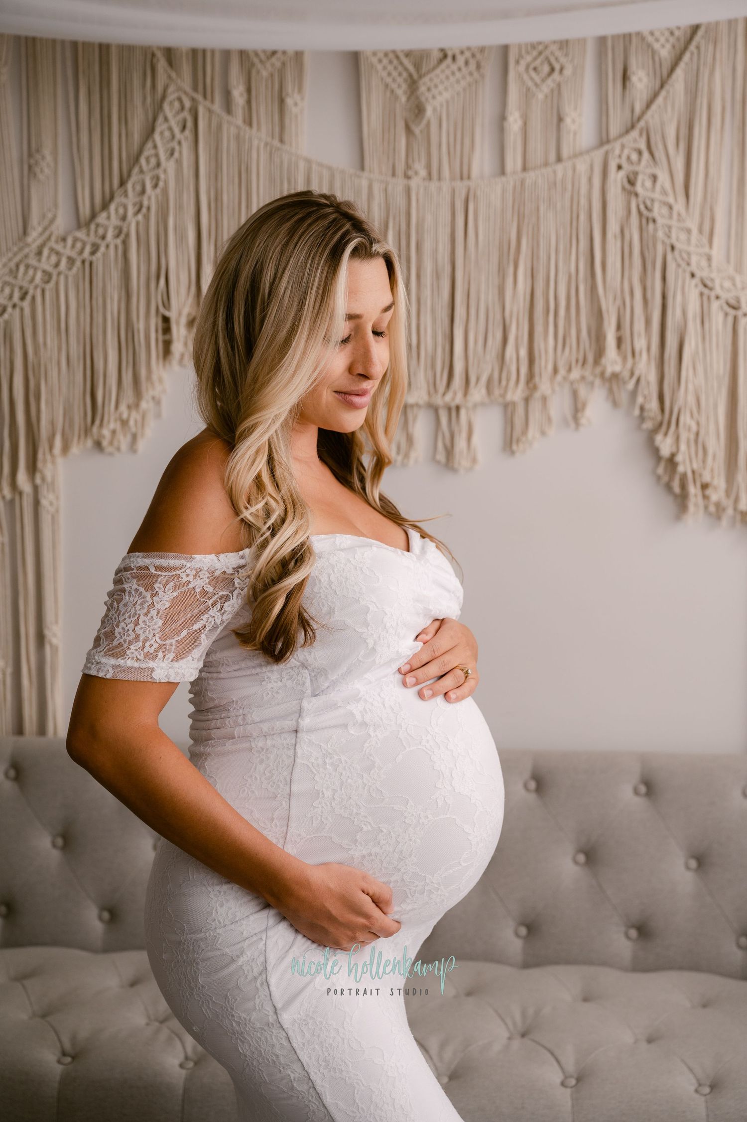 A serene maternity portrait featuring an off-shoulder white lace gown against a macrame backdrop and tufted gray couch.