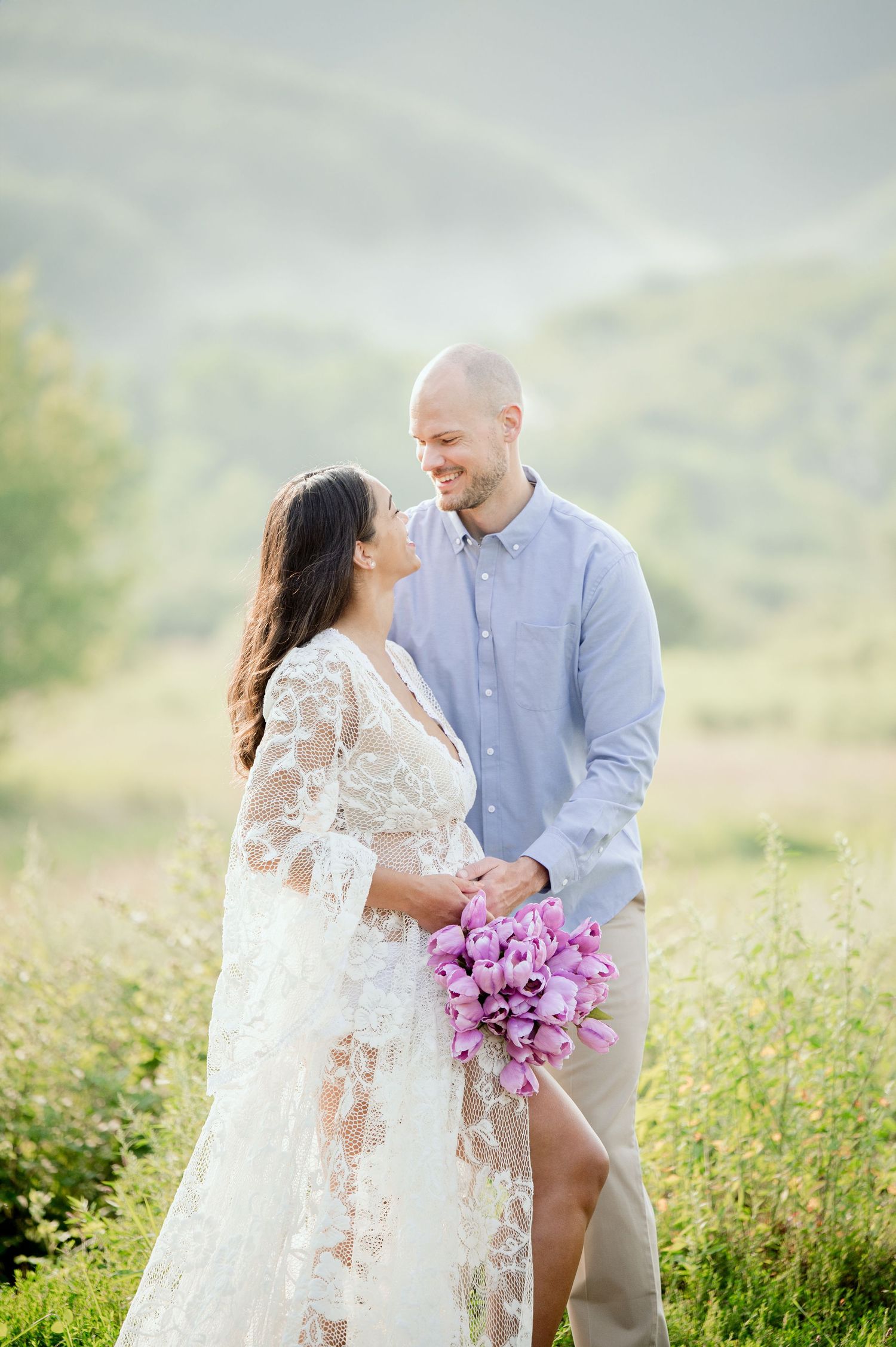 Romantic maternity photo featuring couple in white lace and blue holding purple flowers in a meadow.